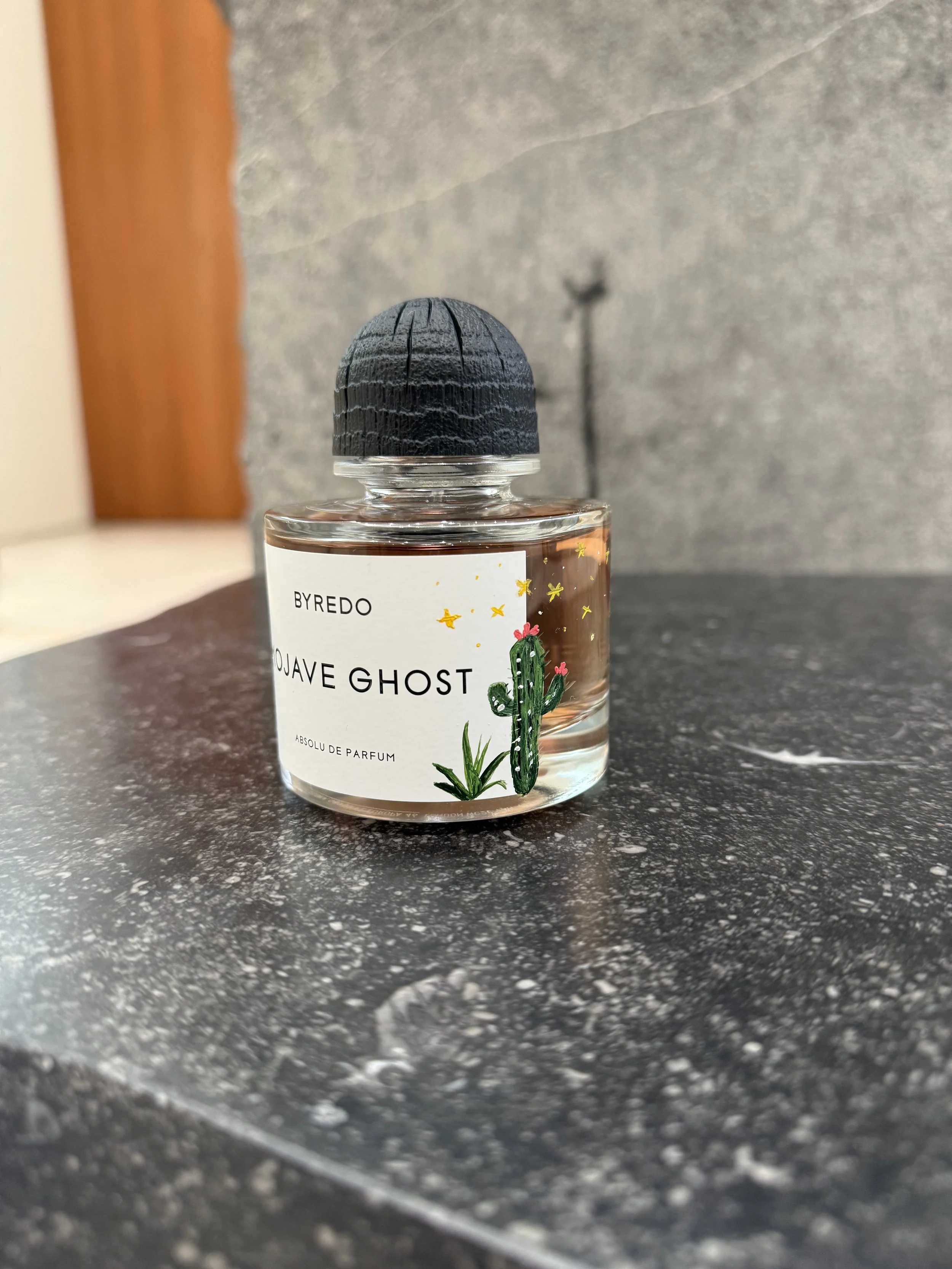 A bottle of Byredo Mojave Ghost perfume with a cactus illustration on the label, placed on a dark marble surface.