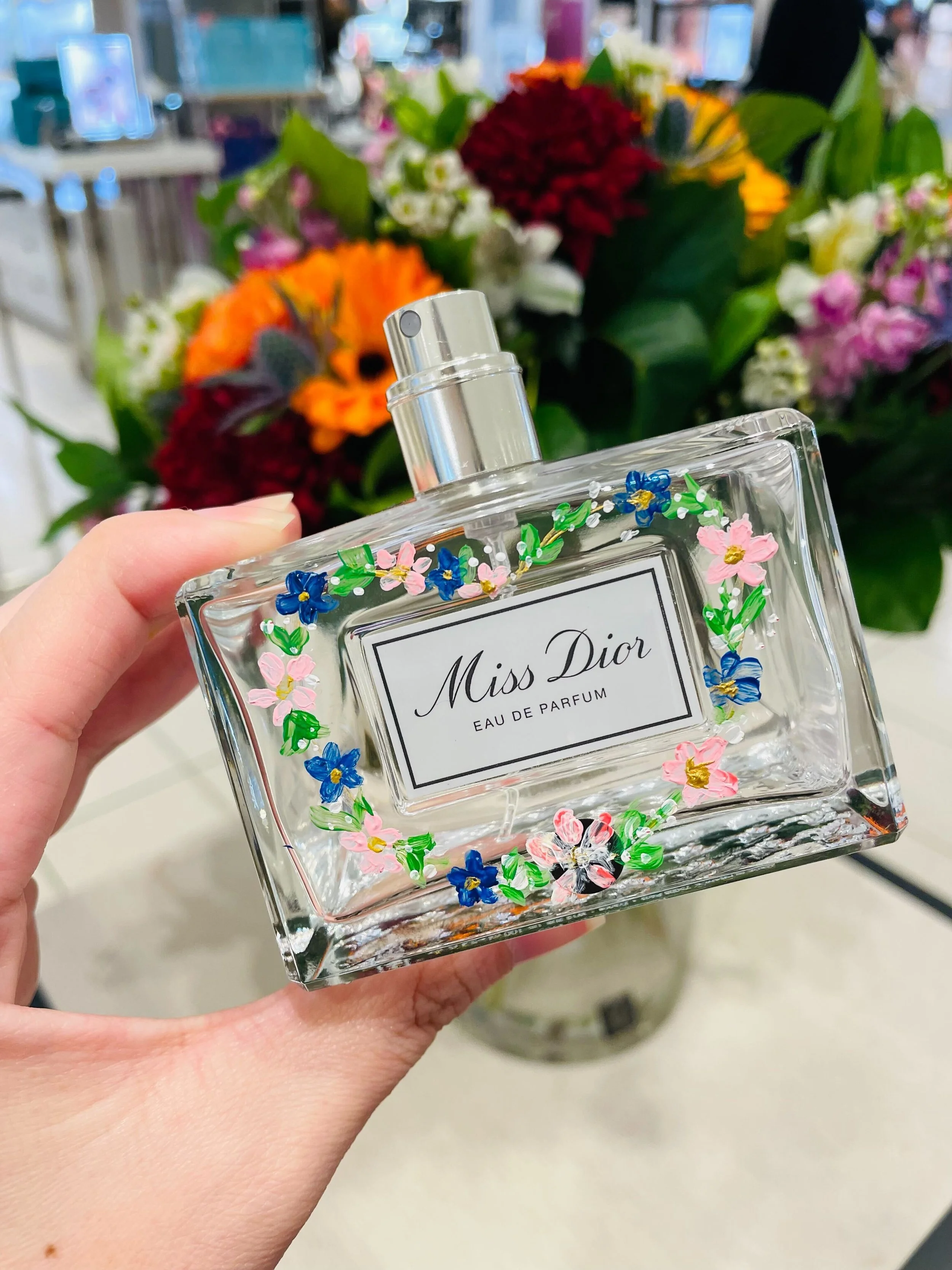 A hand holding a Miss Dior Eau de Parfum perfume bottle with colorful floral decorations on it, in front of a bouquet of mixed flowers.