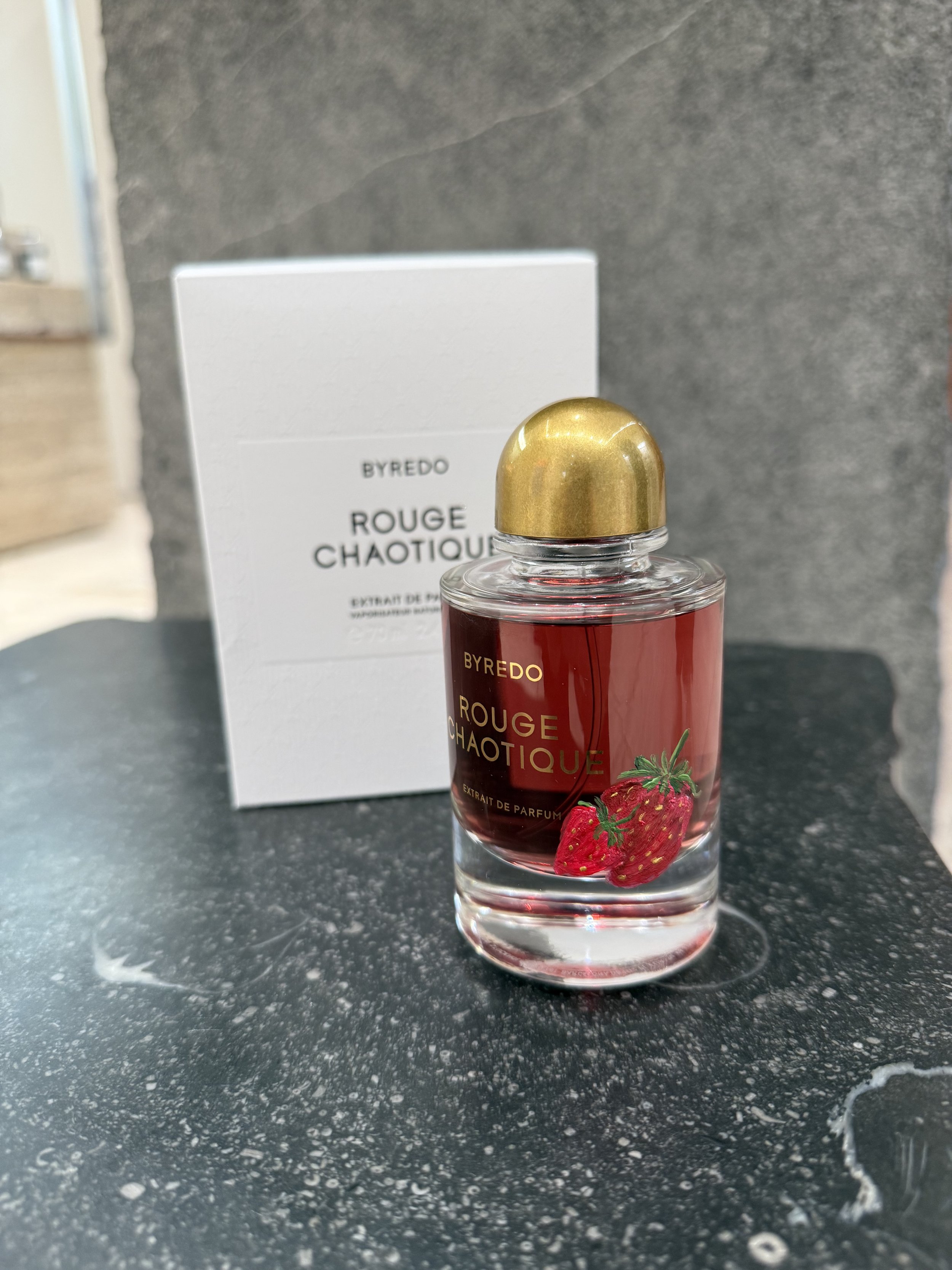 Bottle of Byredo Rouge Chaotique perfume with a strawberry hand-painted on the label, placed in front of its white box packaging on a black speckled surface.