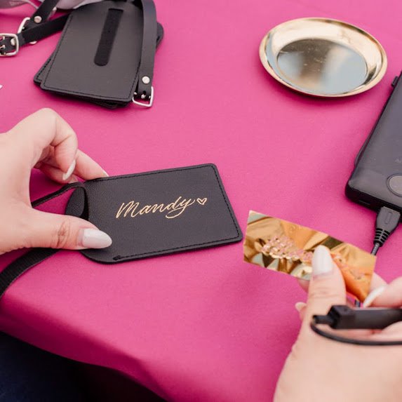A hand holding a name tag with the name 'Mandy' written on it, surrounded by a mirror, a phone, and small pouches on a pink surface.