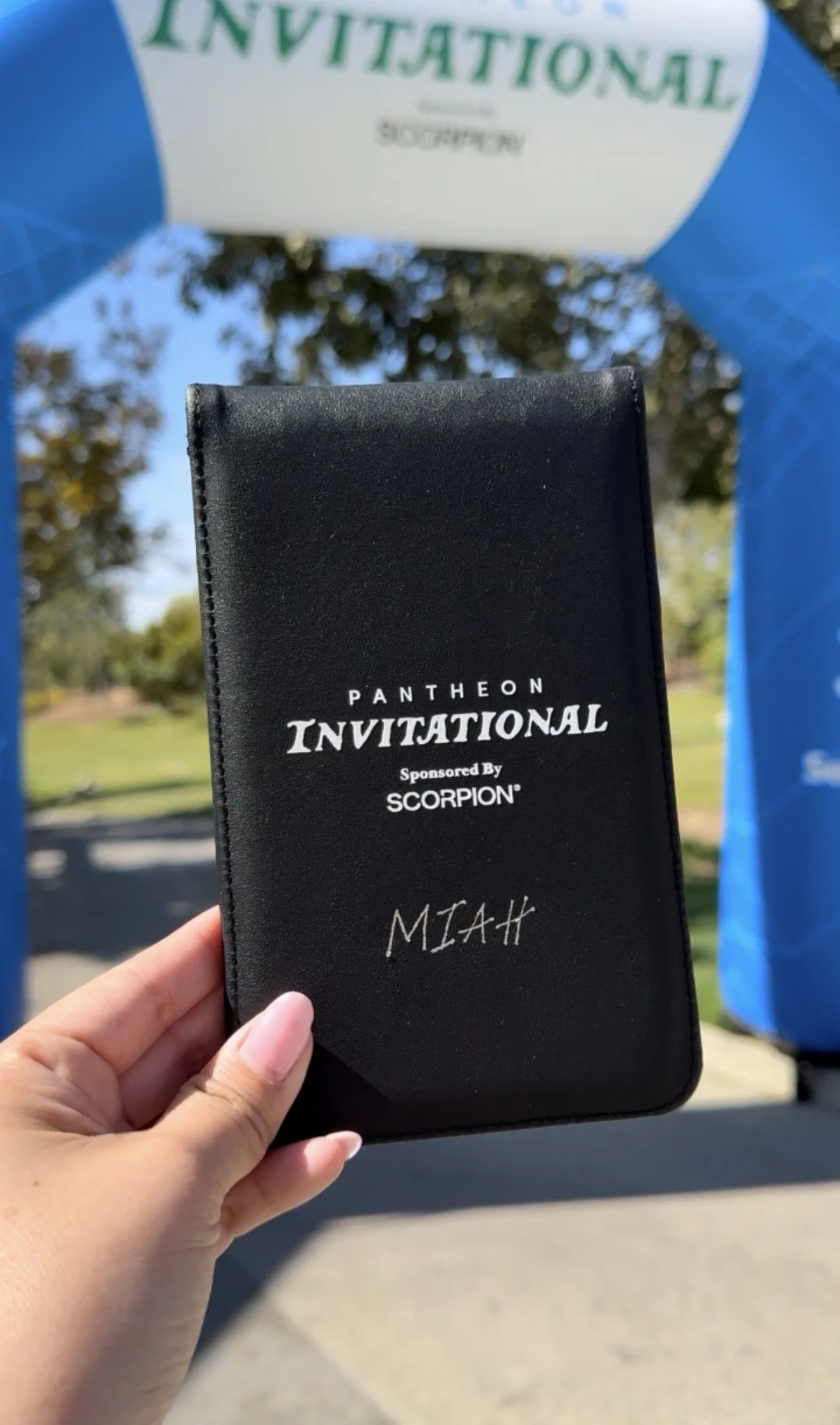 Black leather booklet with white text that reads 'Pantheon Invitational Sponsored By Scorpion Miah,' held by a person's hand in front of a blue archway at an outdoor event.