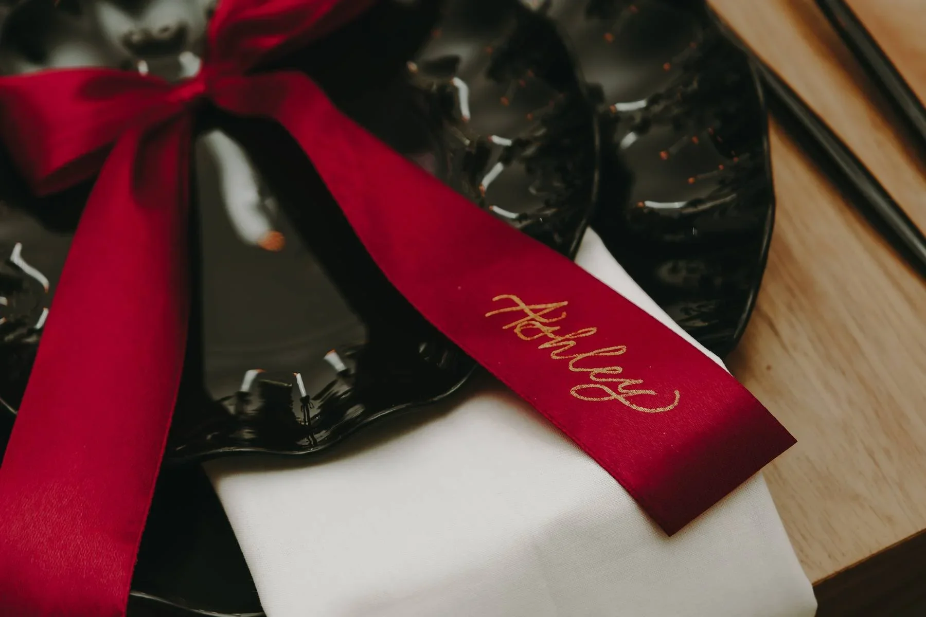 Black ceramic Christmas wreath with holly accents, decorated with a red ribbon and a small white gift box, on a white fabric surface, next to a wooden table.