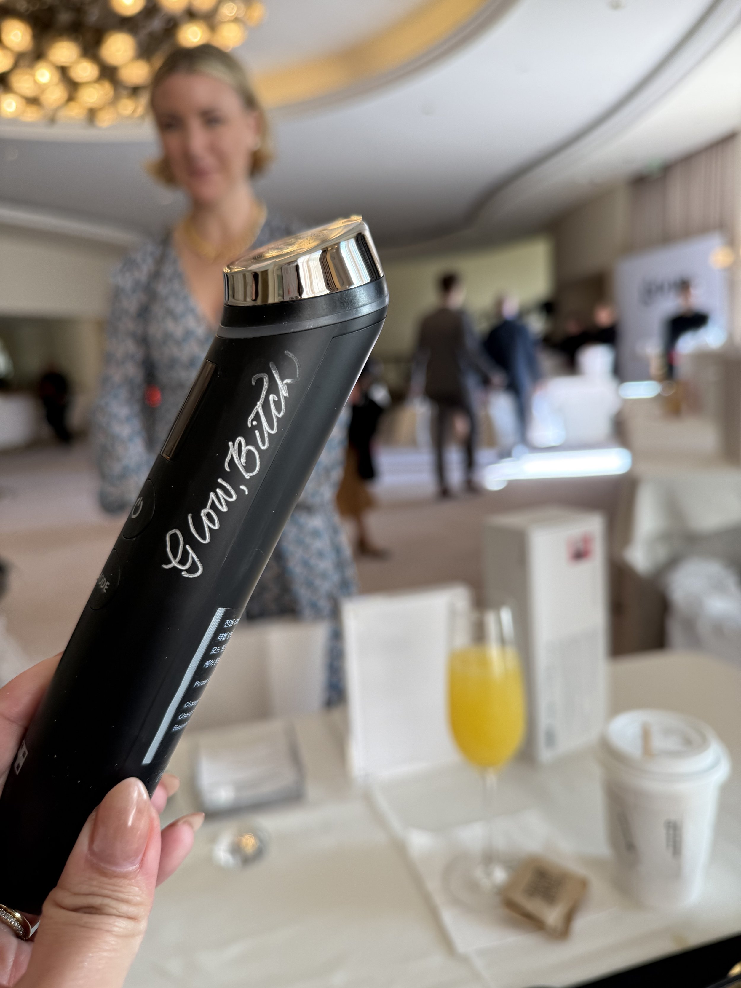 Close-up of a black tumbler with white writing, held in the foreground at a social gathering, with a blurred background showing a woman, people, and a table with drinks and food.