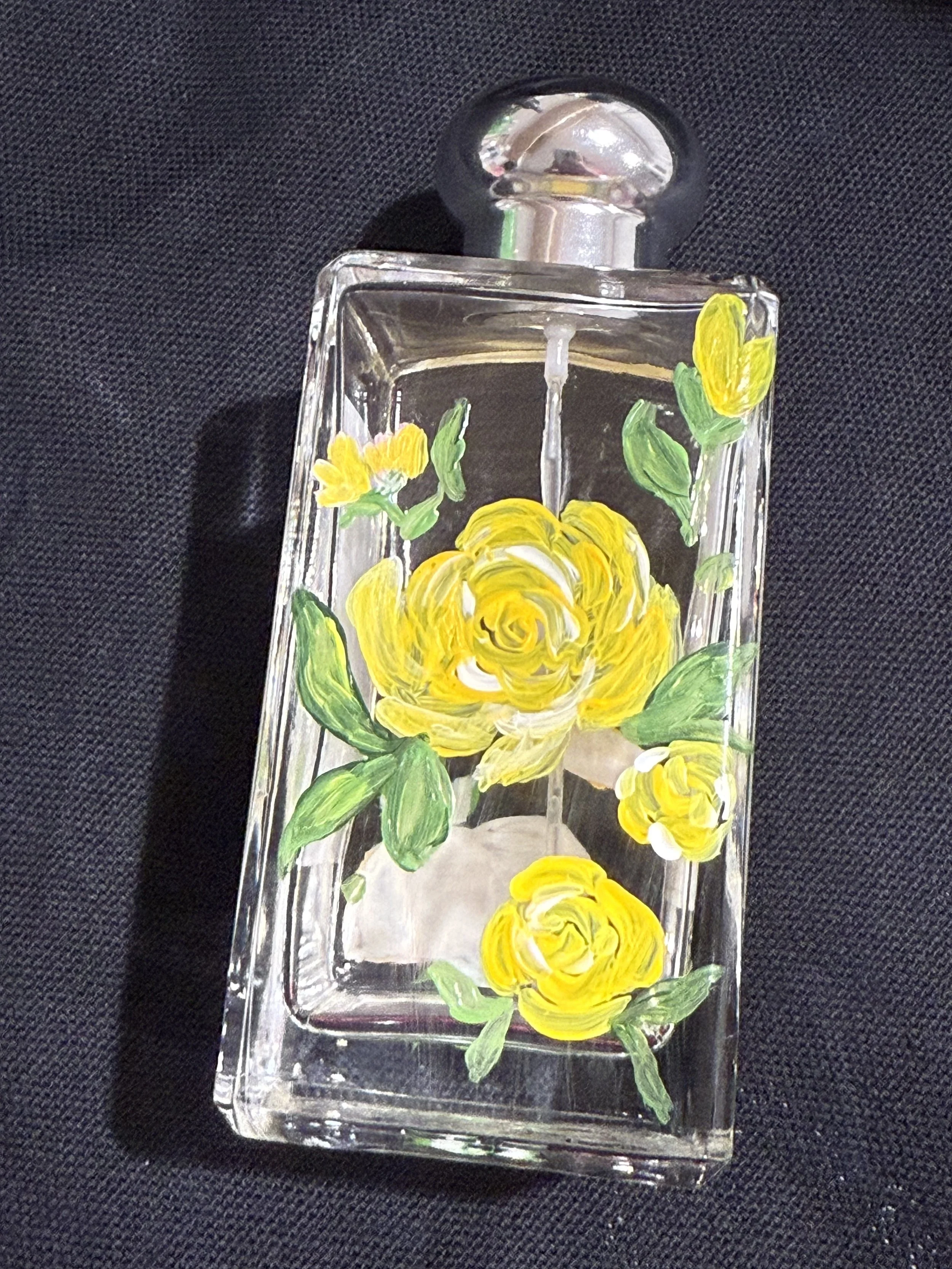 A glass perfume bottle with a silver cap, decorated with hand-painted yellow roses and green leaves, placed on a dark fabric background.