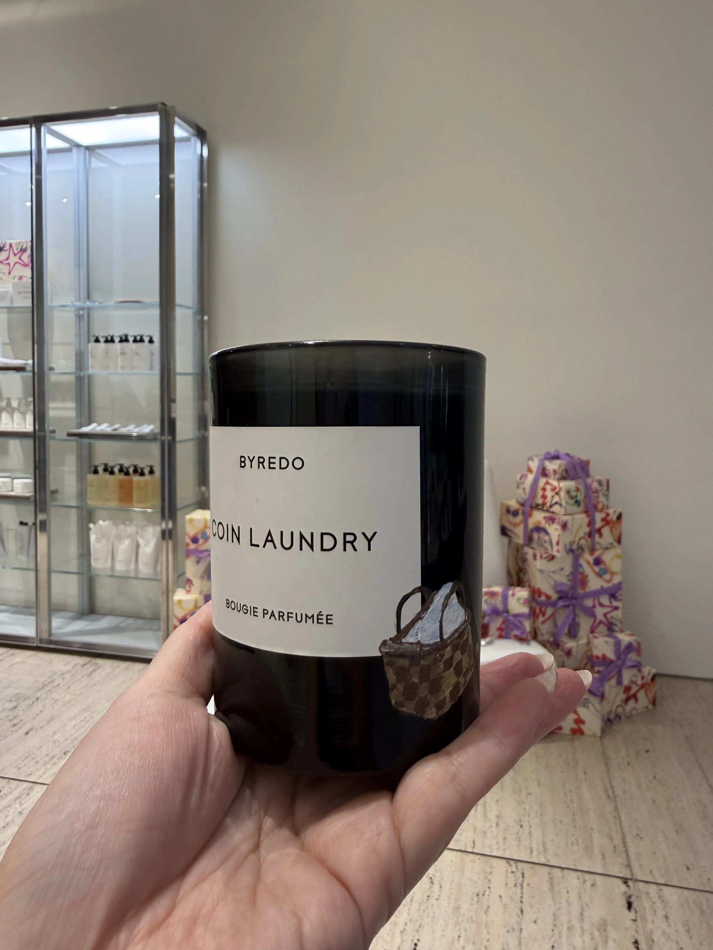 Black candle labeled 'Byredo Coin Laundry Bougie Parfumée' held in hand, with wrapped gifts in the background.