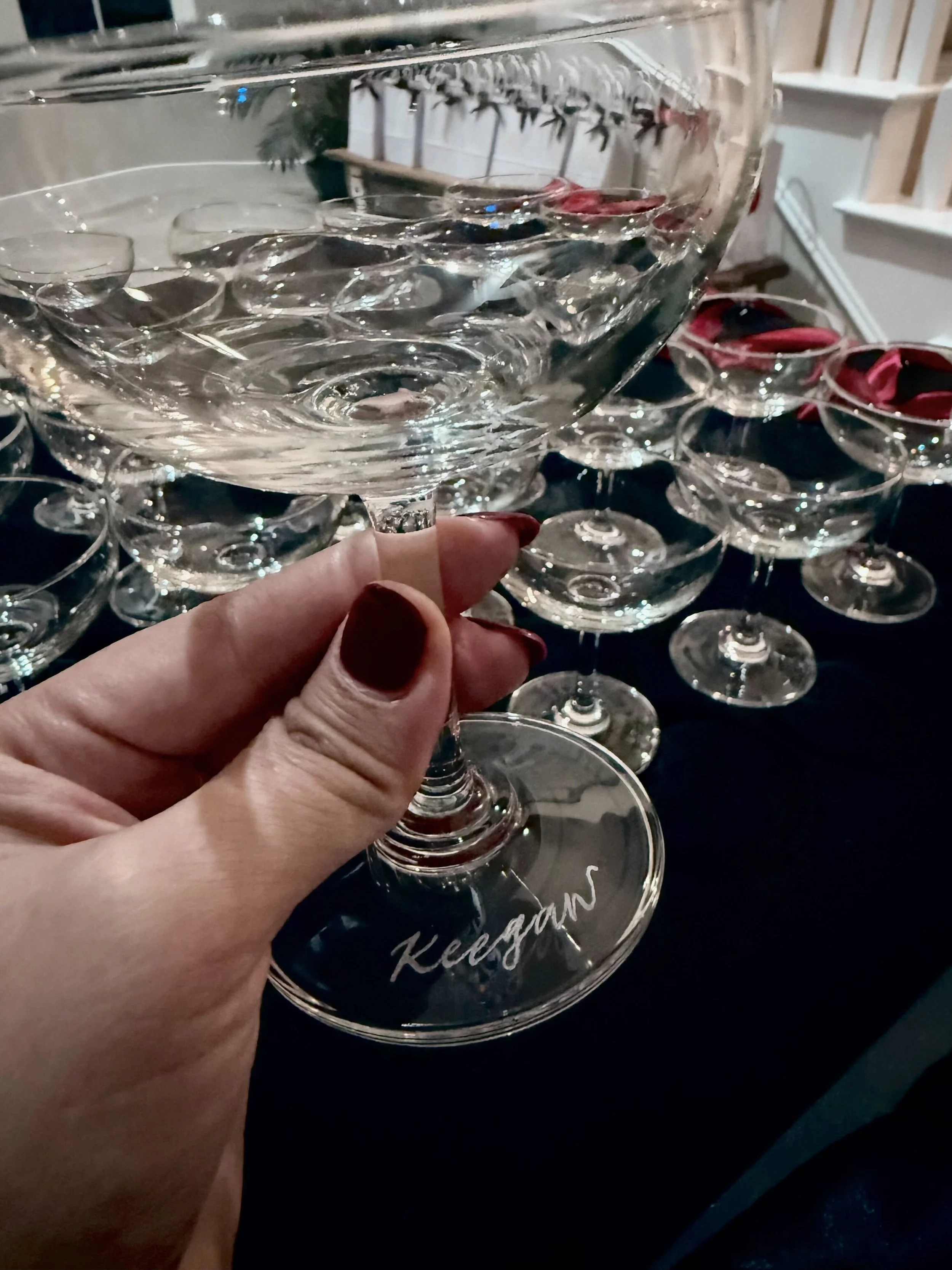Close-up of a hand with dark red nail polish holding a clear glass champagne coupe glass. The glass is empty. In the background, there are many more empty coupe glasses arranged on a dark table.