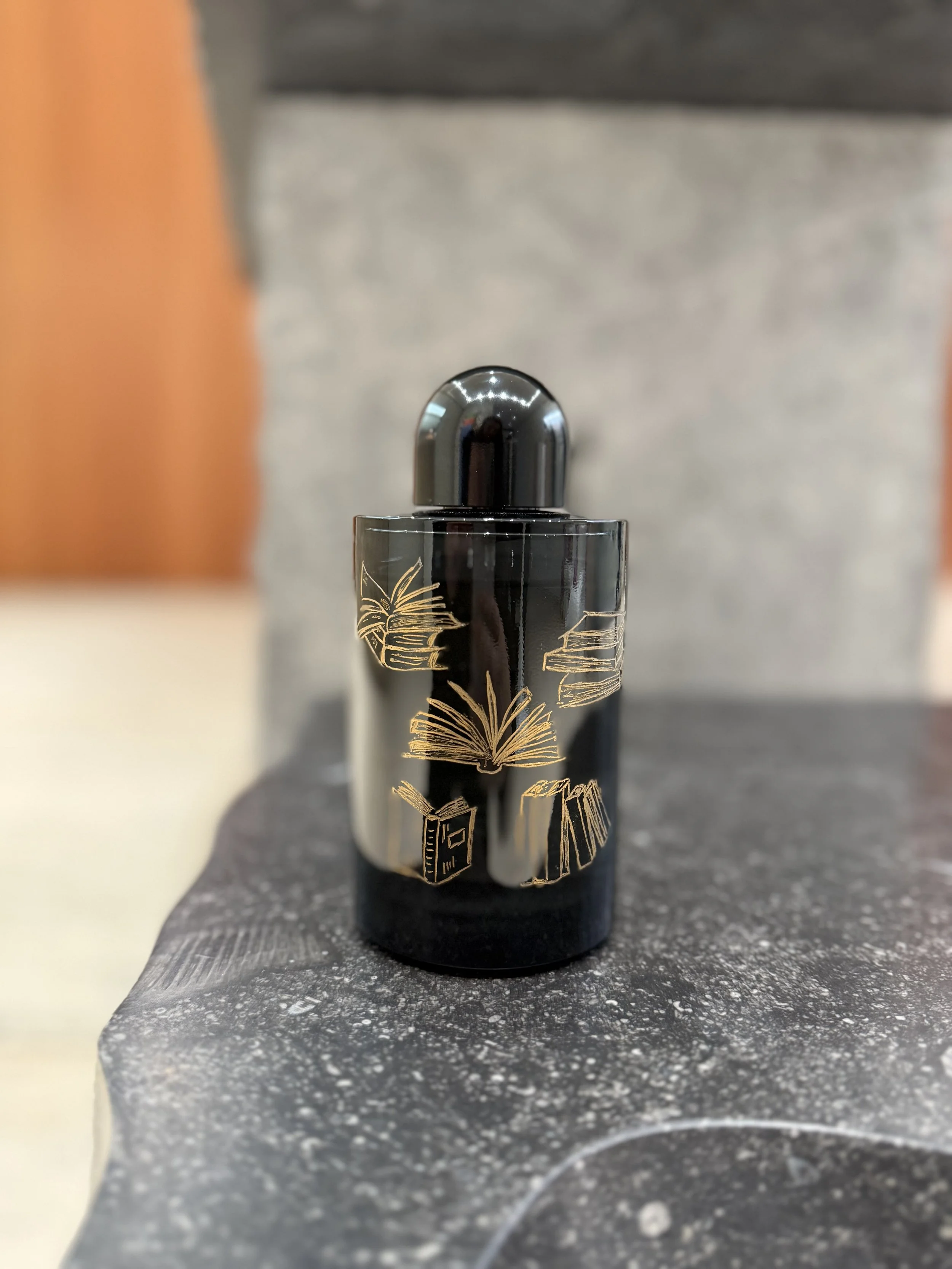 Black perfume bottle with gold book and paper designs on the front, placed on a dark, textured surface.