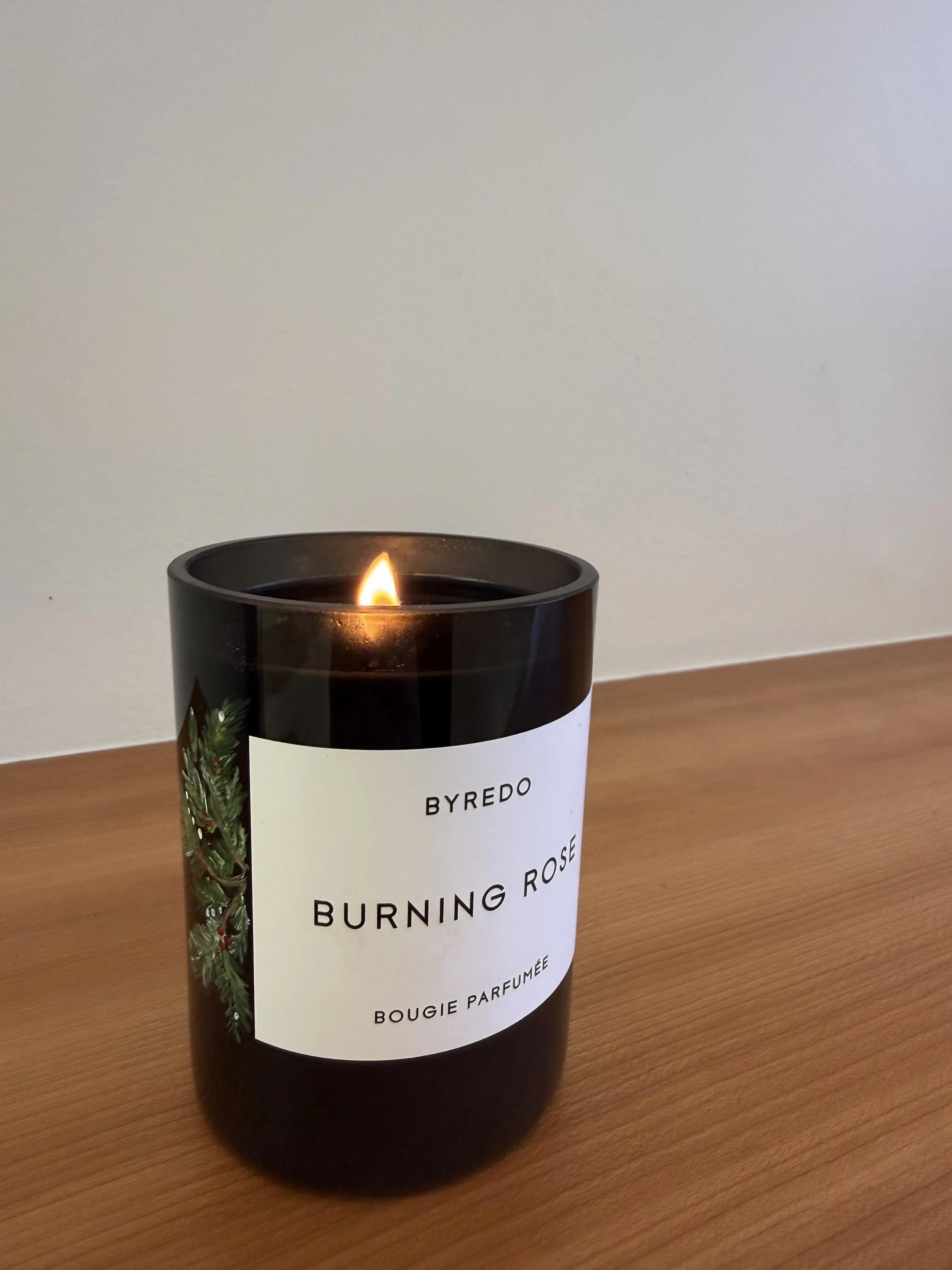 Black scented candle labeled 'Byredo Burning Rose Bougie Parfumée' on a wooden surface with a small flame