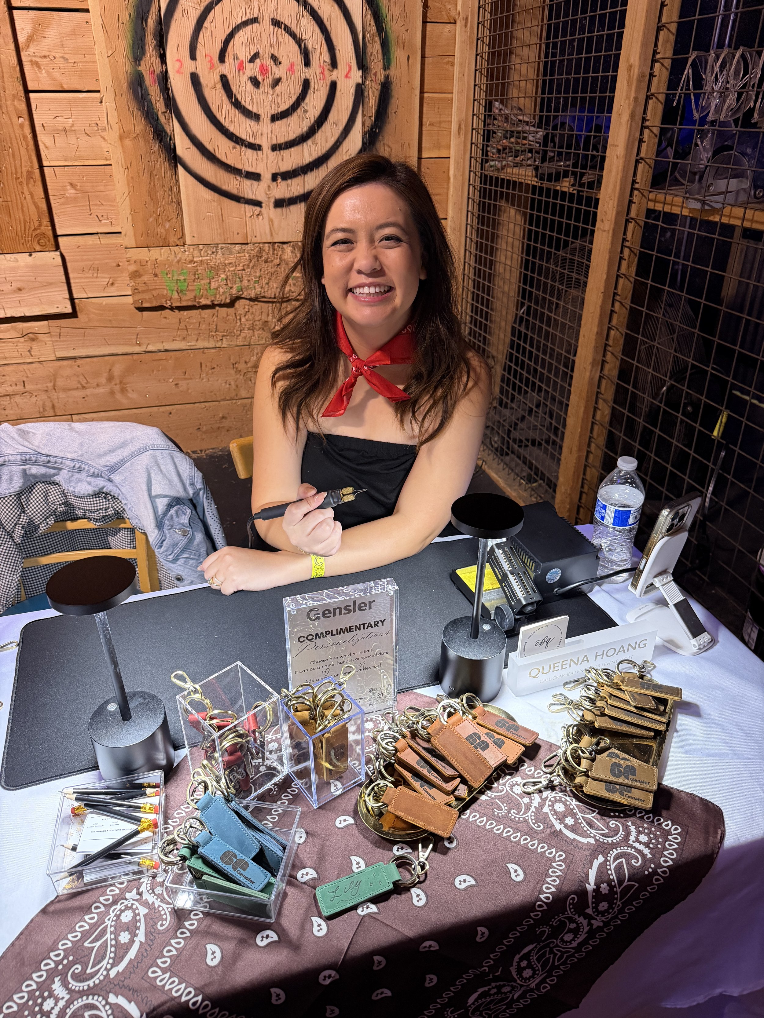 Smiling woman with shoulder-length brown hair sitting at a table with promotional keychains, pens, and business cards. She has a red bandana around her neck and holds a pen. The background features a wooden wall with a target painted on it and some c