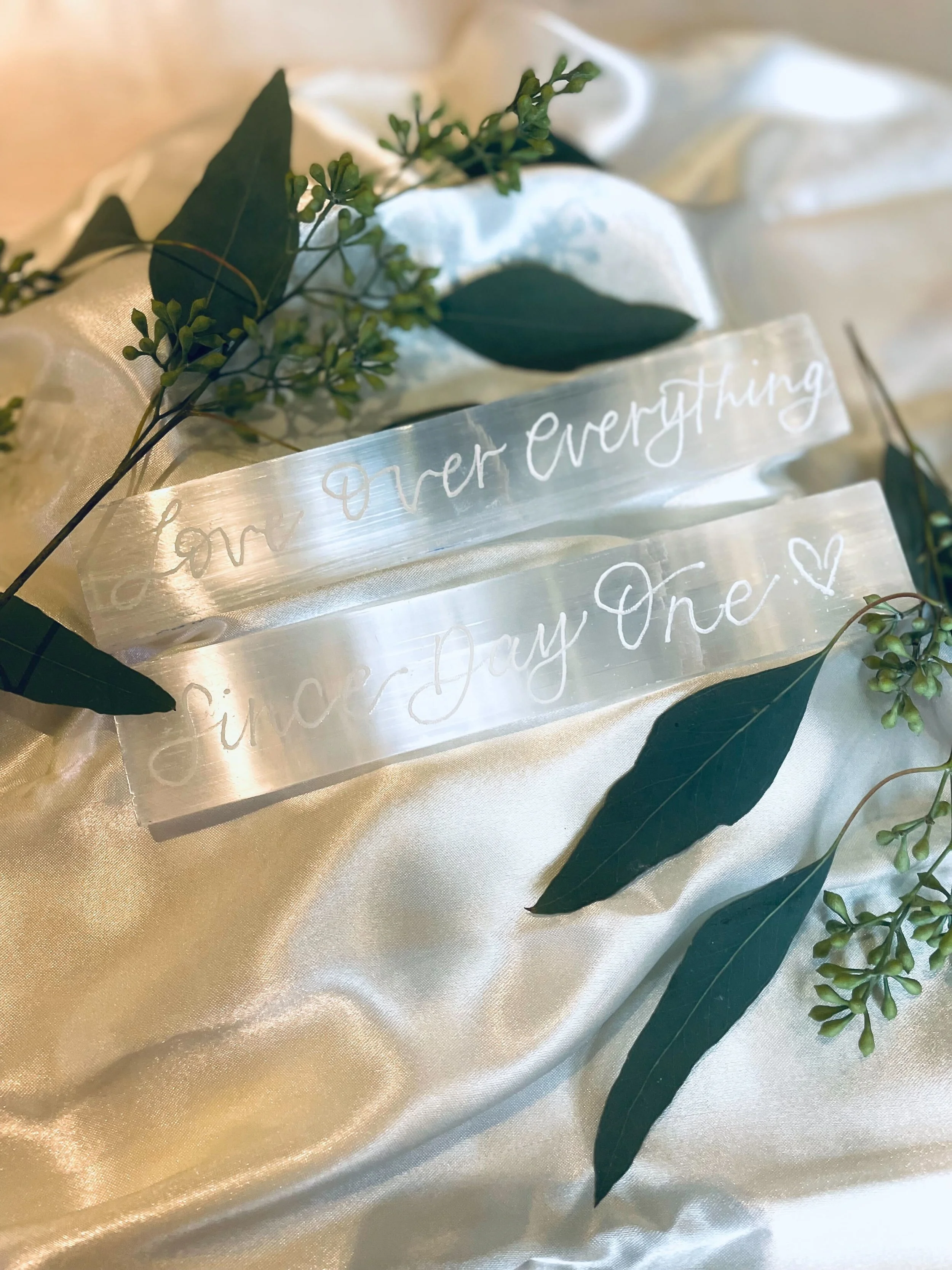A clear, engraved acrylic sign reading "Love Ever Everything" and "Forever And One" with a small heart, placed on satin fabric and decorated with green leaves and small green buds.