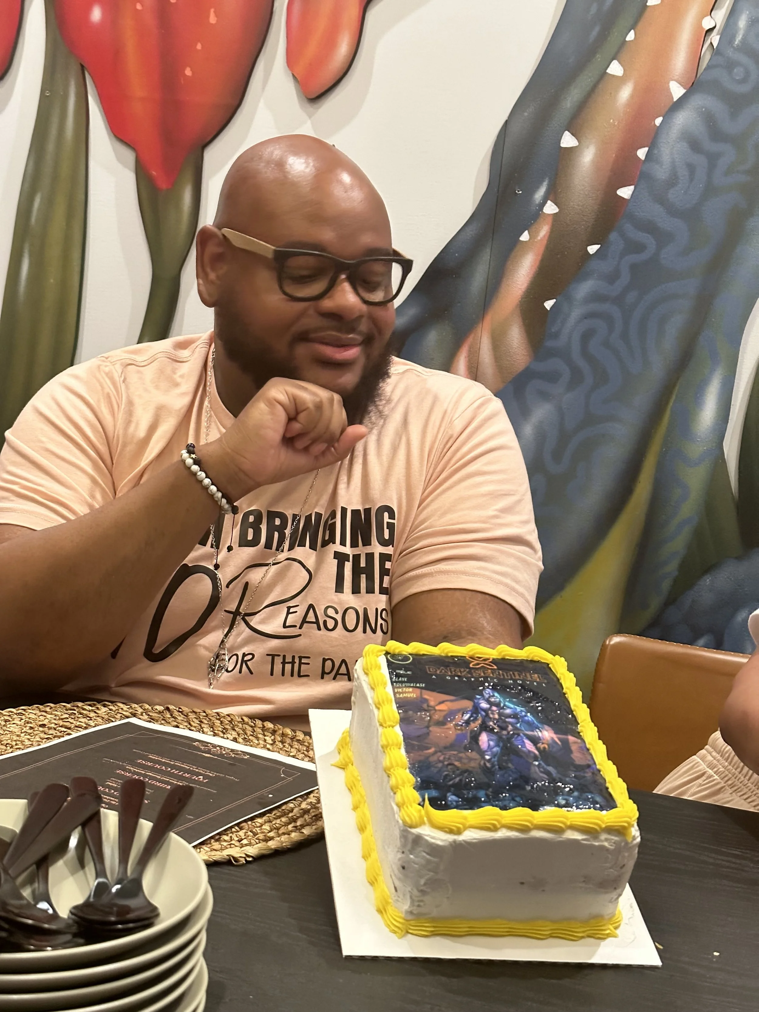 Man wearing glasses and a peach-colored shirt sits at a table with a rectangular cake decorated with a printed image on top. The cake has a yellow border, and there are plates and utensils nearby. Artistic wall decor is partially visible in the background.