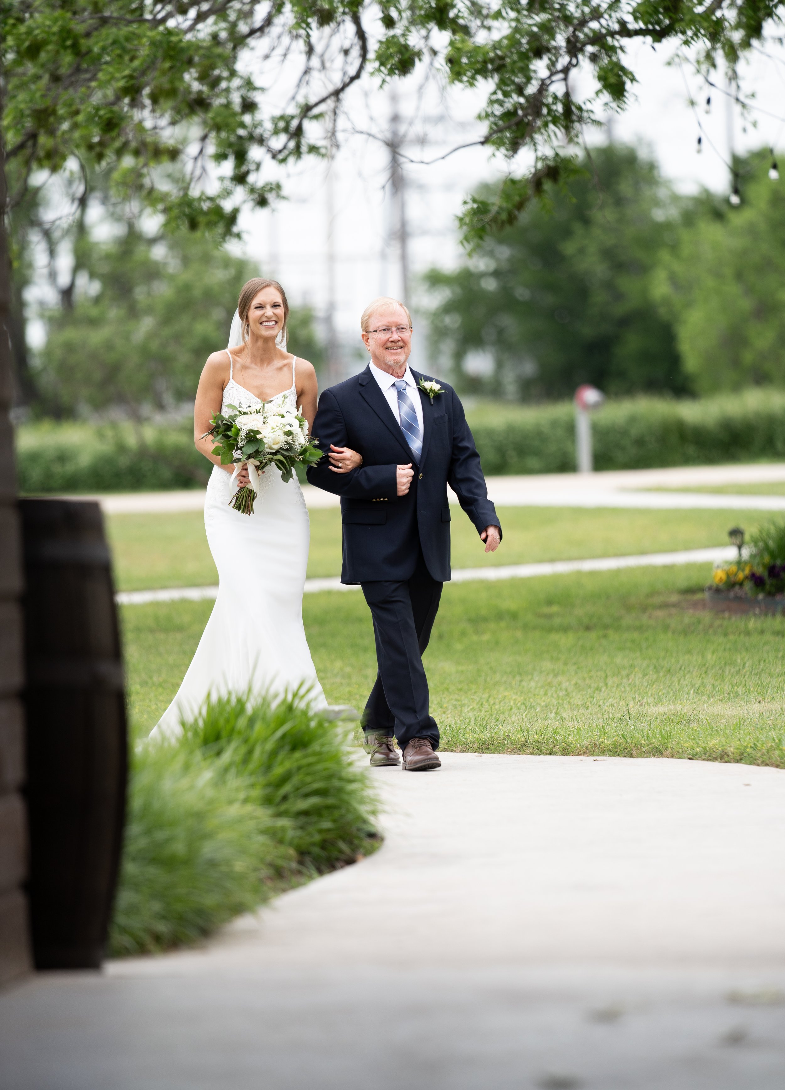 A bride walking down the aisle with her father, holding a bouquet of white flowers and smiling, outdoors on a cloudy day with green trees and grass in the background.