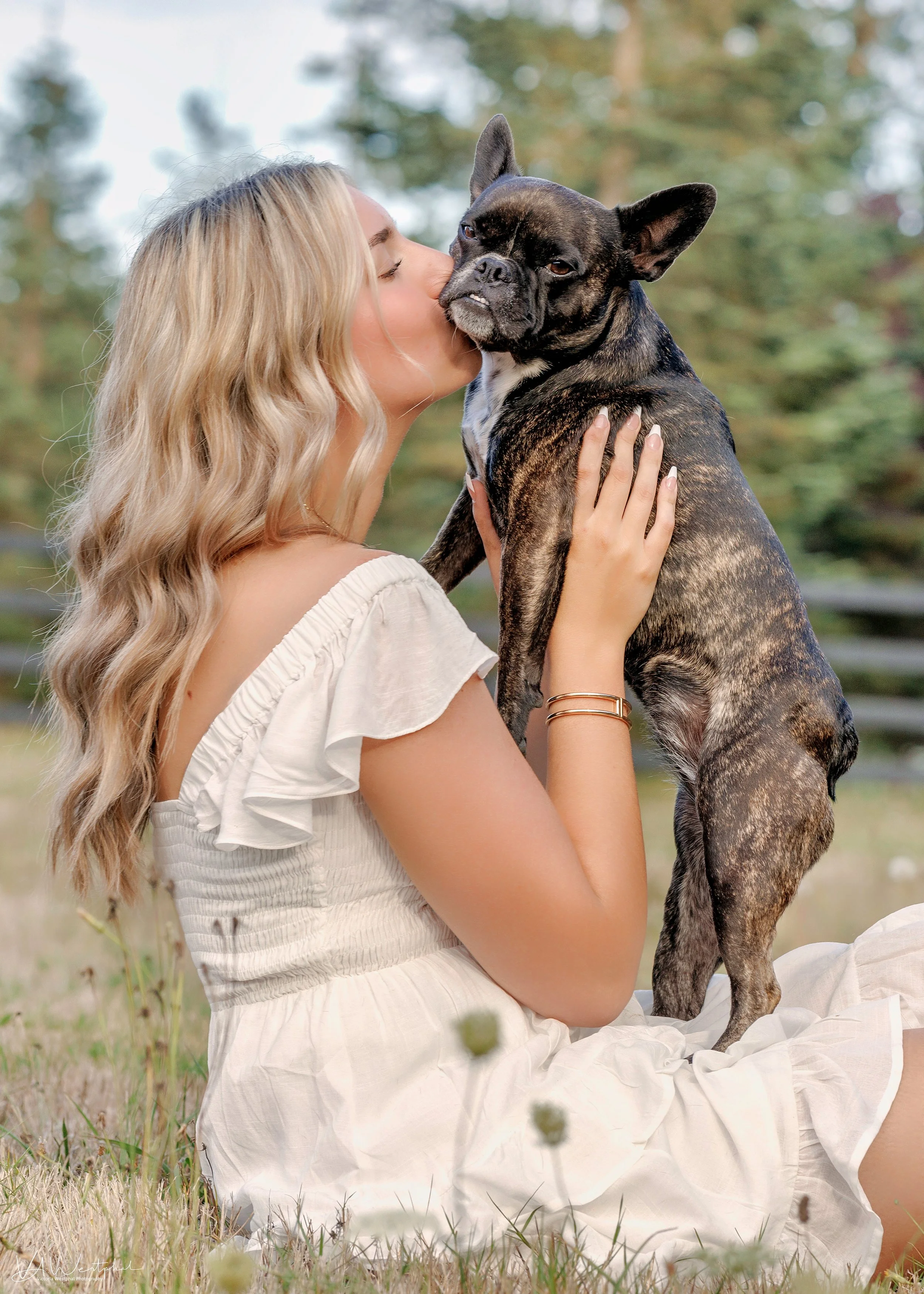 A woman with blonde hair, wearing a white dress, sitting on the grass outdoors, holding and kissing a brindle French Bulldog.