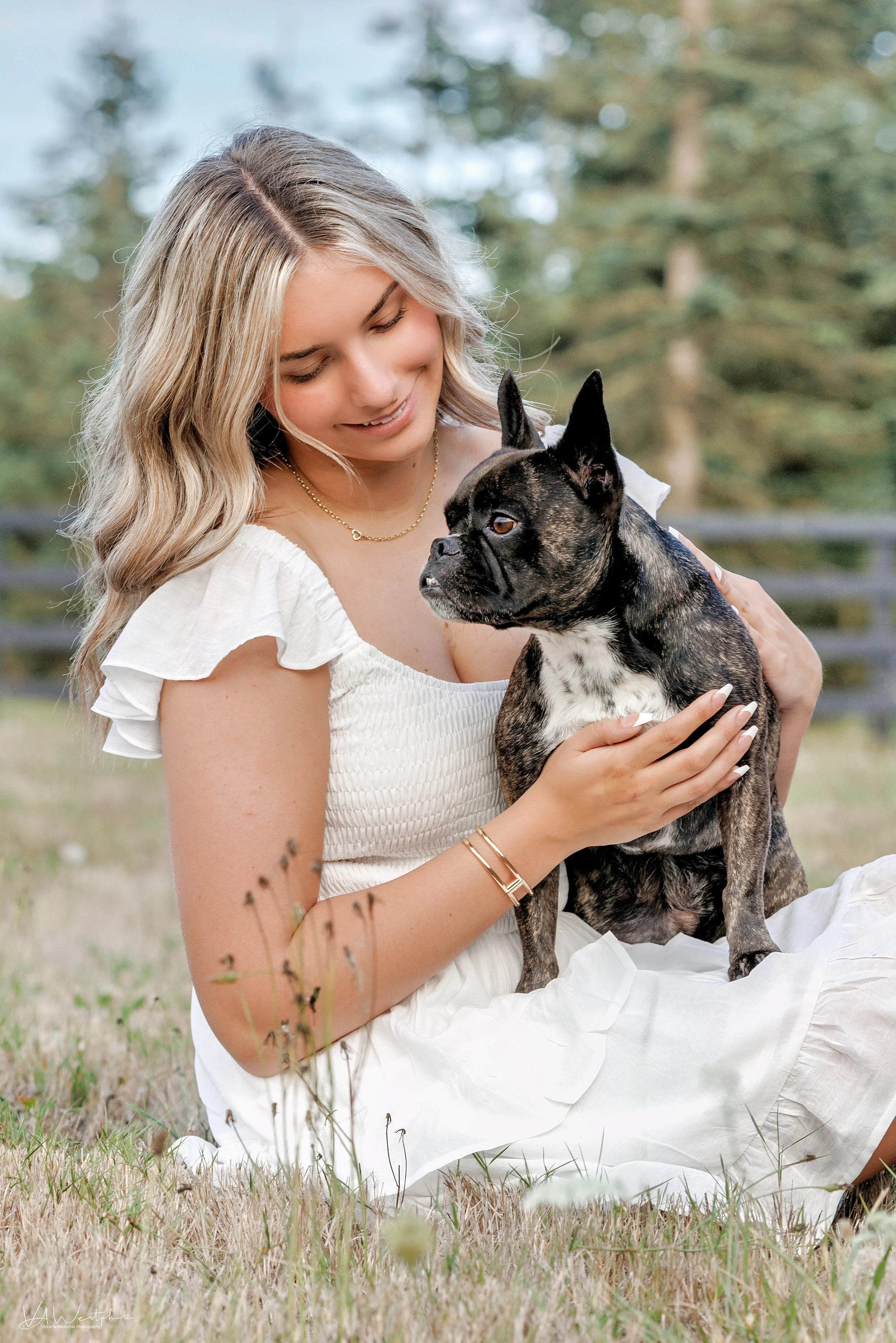 A young woman with blonde hair sitting on grass outdoors, holding and looking at a small brindle French Bulldog.
