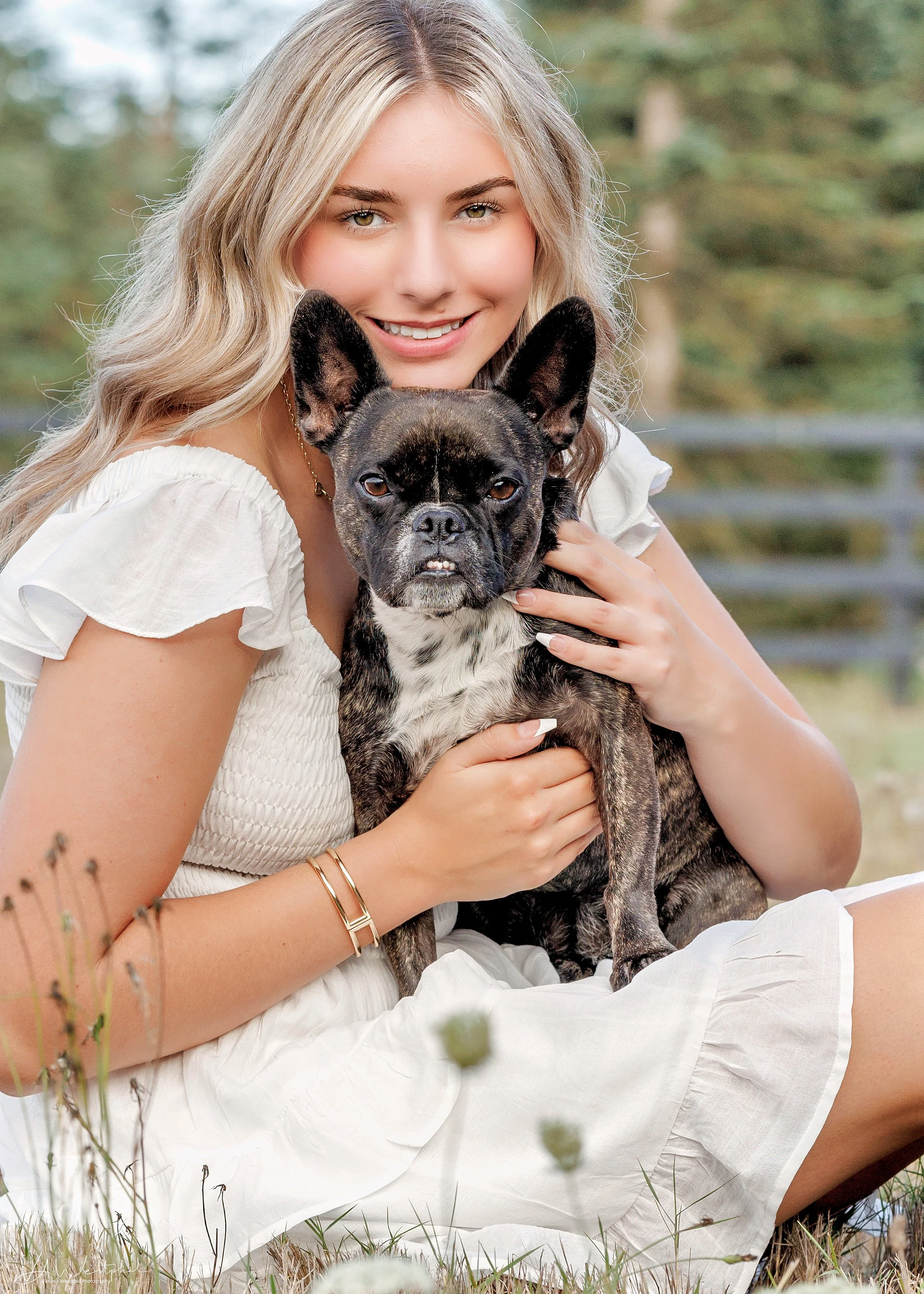 A young woman with long blonde hair smiling and holding a brindle French Bulldog outdoors in a grassy area.