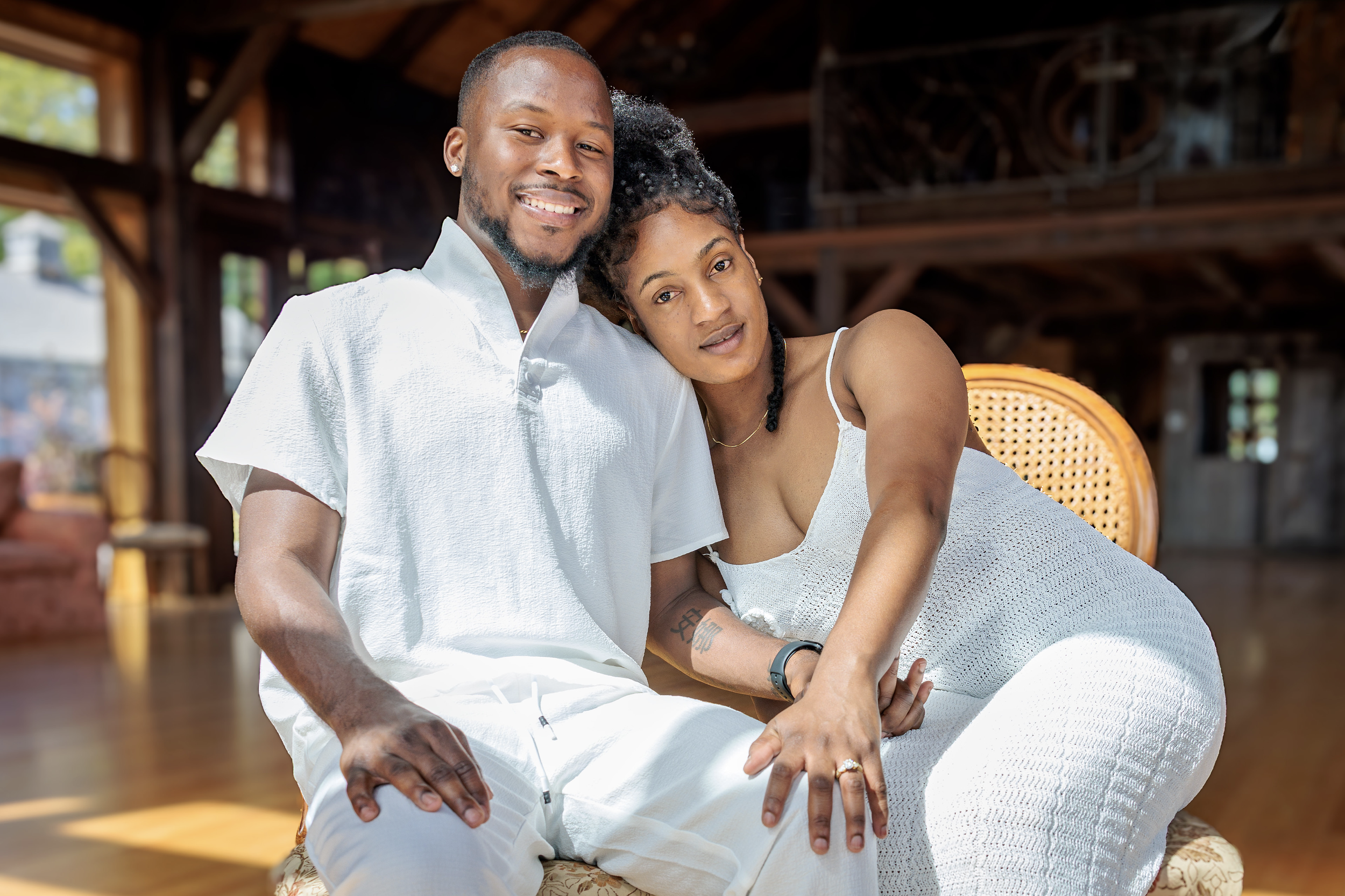 A smiling couple, dressed in white, sitting close together in a warmly lit, wooden interior.