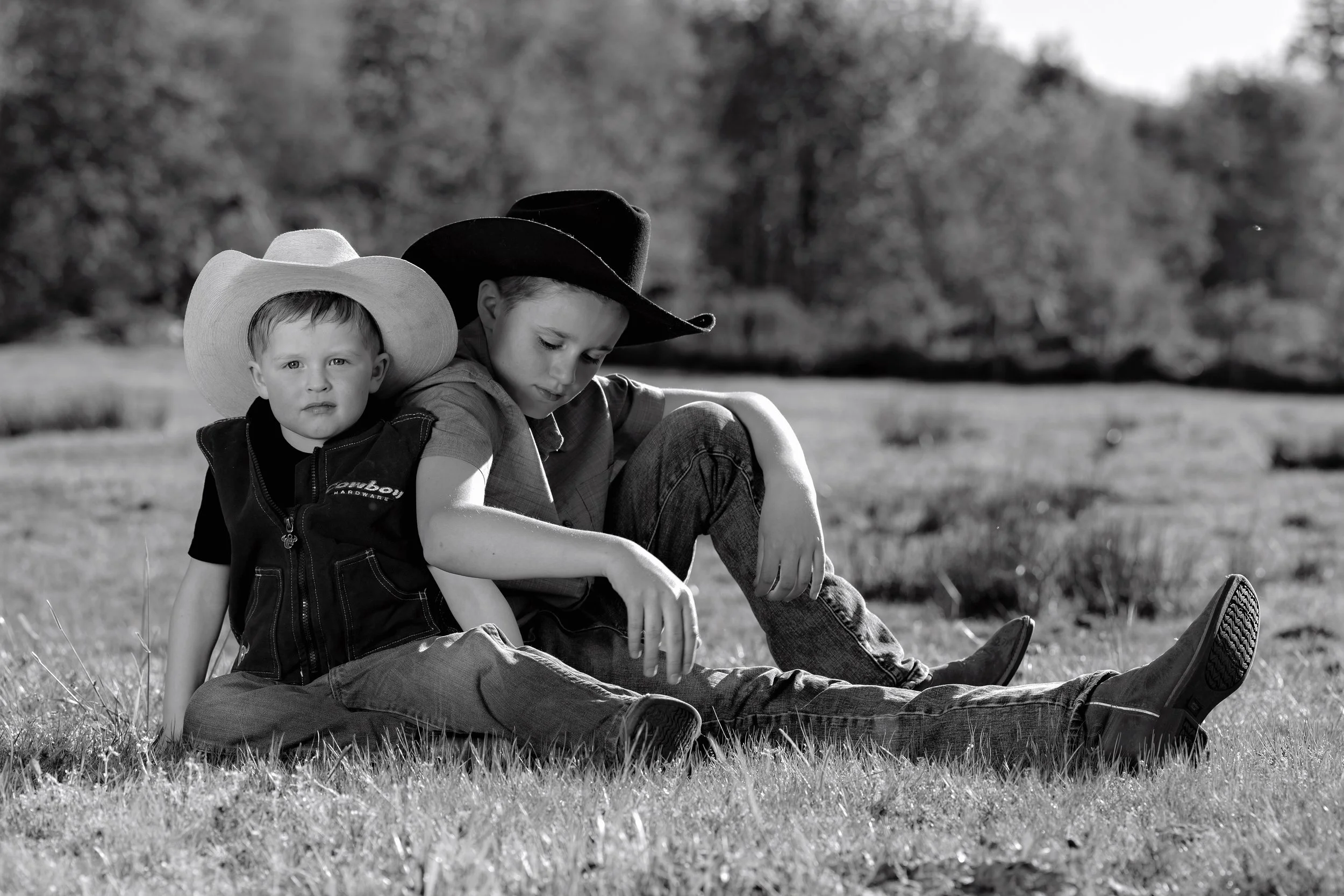 Two young boys sitting on grass outdoors, wearing cowboy hats. The older boy is looking down, while the younger boy is sitting behind him, looking forward. The background shows trees and an open field.