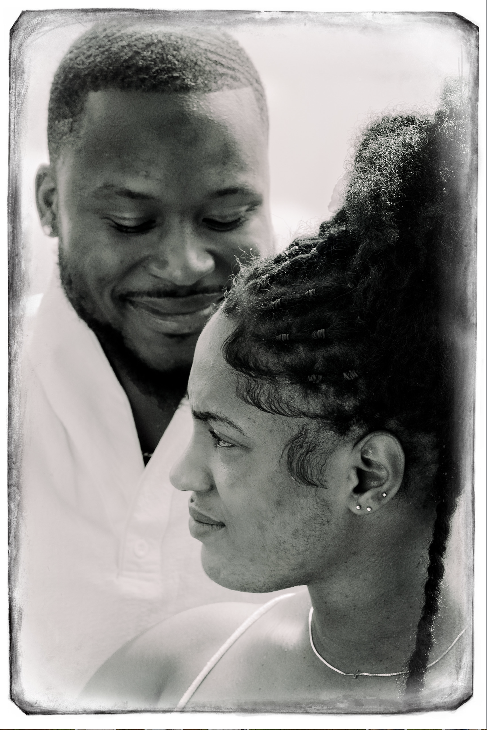 A black-and-white close-up photo of a smiling man looking at a woman with an elaborate hairstyle, both viewed in profile.