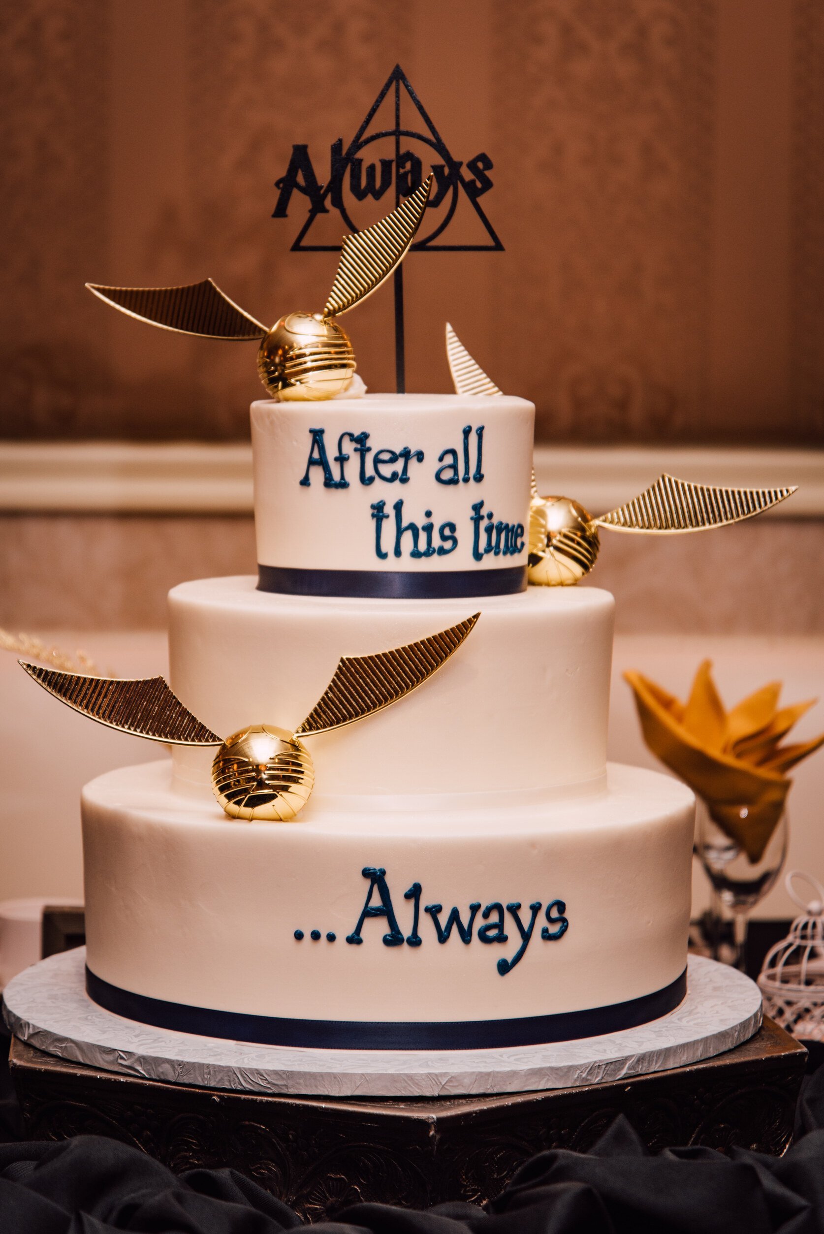  a harry potter wedding cake sits on a table decked out with the always symbol and quidditch snitch with the quote “after all this time… always” piped on the side of the 3 layer cake 