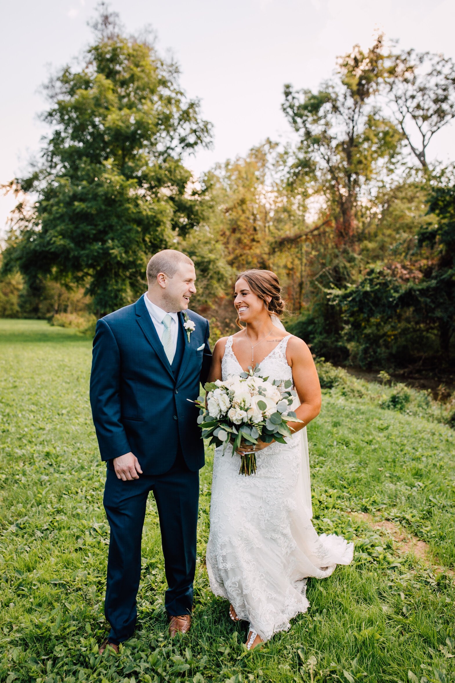  The bride and groom stand in a field smiling at each other while the bride holds her white and green bouquet at their barn wedding 