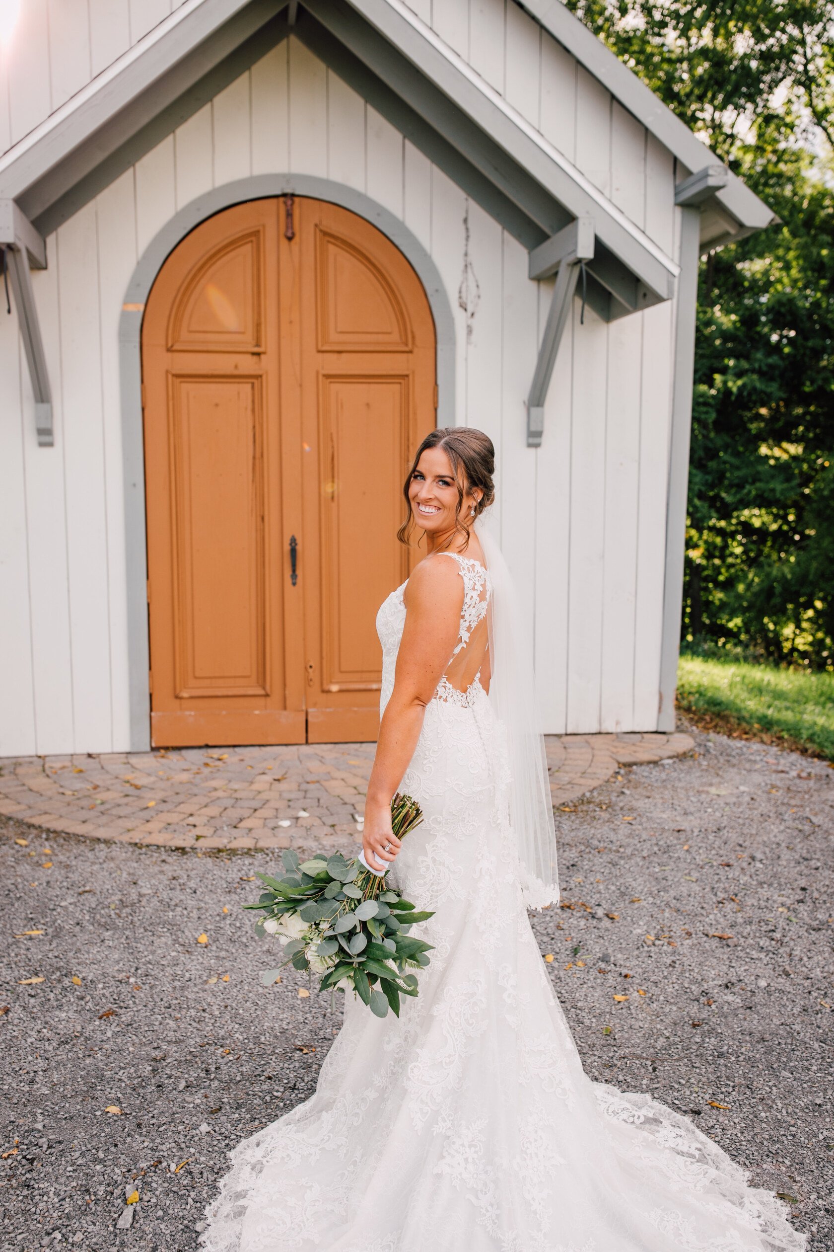  The bride smiles as she looks over her shoulder while holding her bouquet of flowers and standing in front of a chapel at hayloft on the arch 