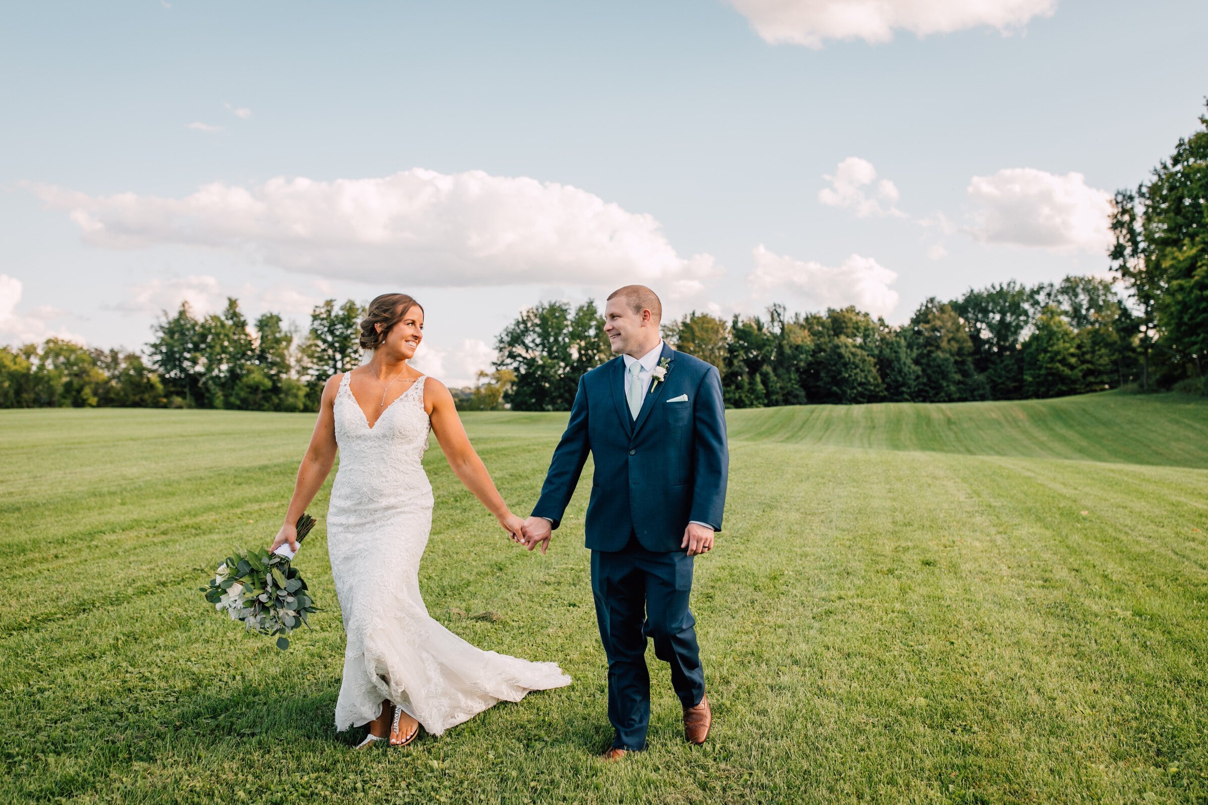  The bride and groom walk hand in hand in a field while looking at each other during their barn wedding 