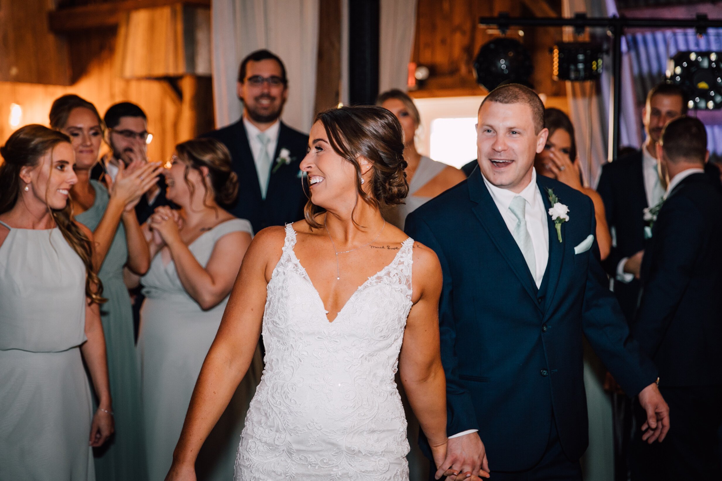  The bride and groom smile as they are entering their rustic barn wedding reception and surrounded by their wedding party 