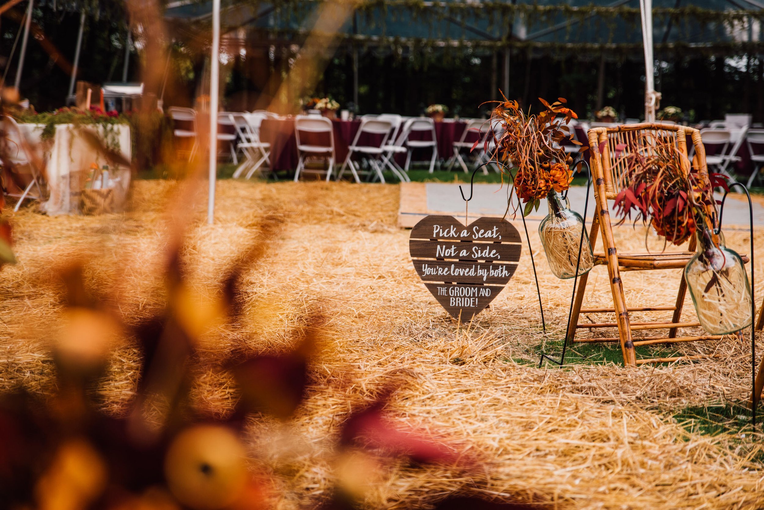  A wood sign that says “Pick a seat, not a side. You are loved by both the groom and bride.” Sits next to a rattan folding chair at an outdoor fall wedding 