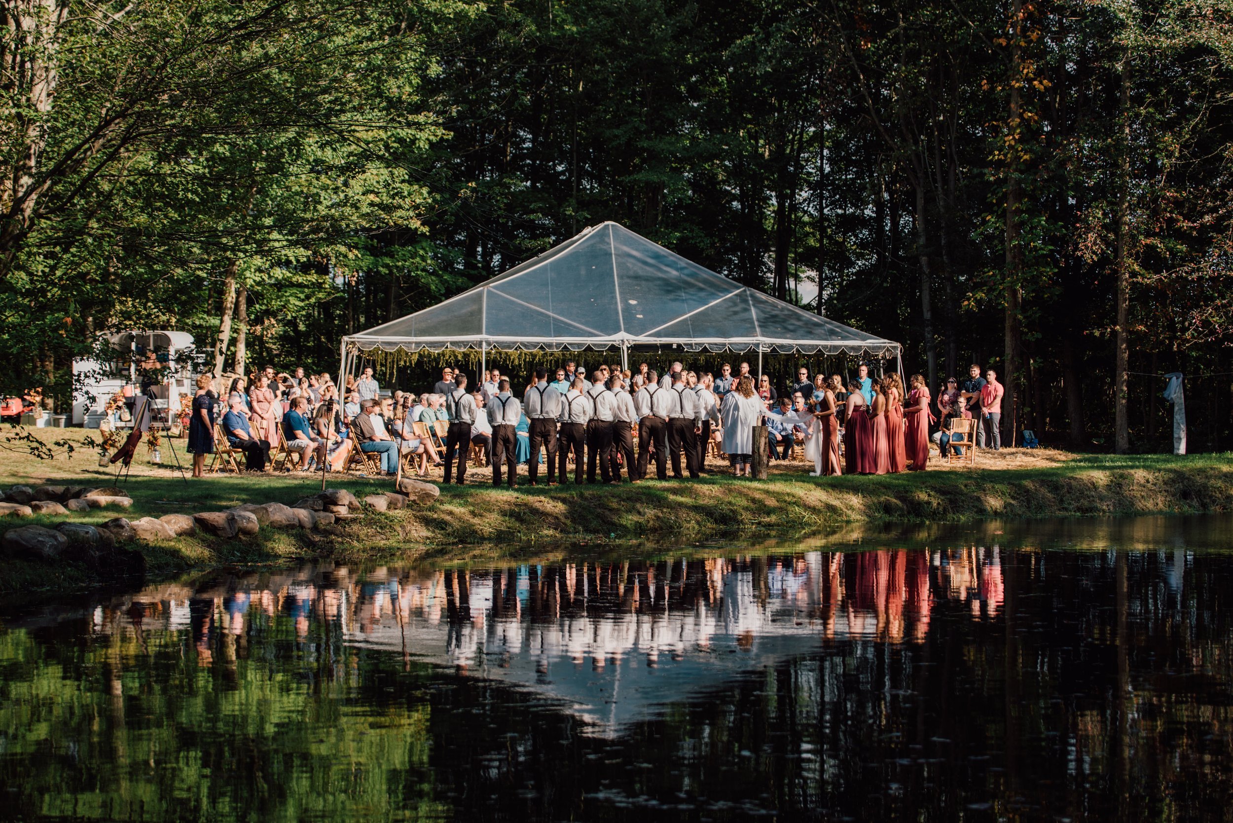  view from behind the outdoor fall wedding ceremony across a pond reflecting the image of the wedding party back  