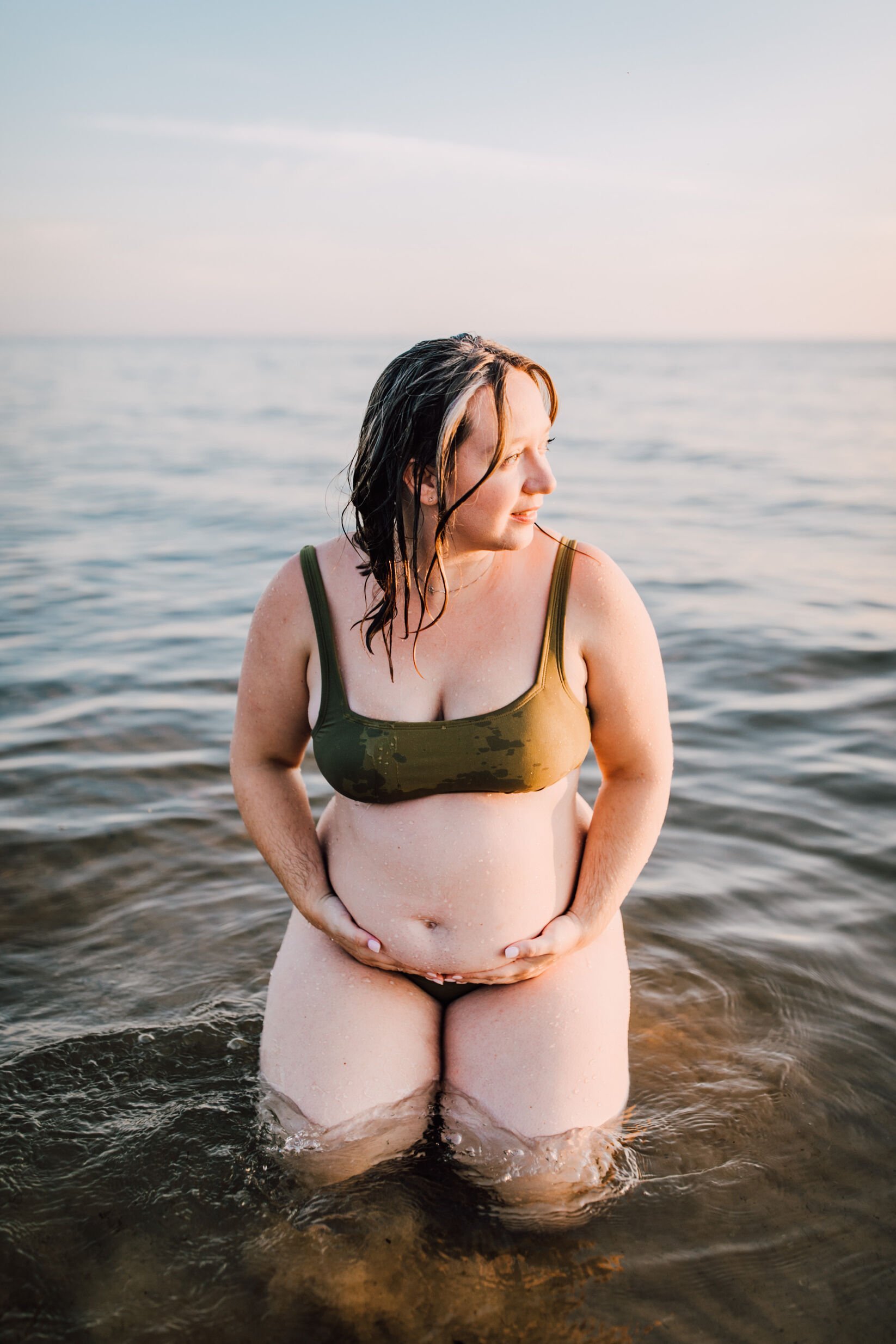  A pregnant woman with wet hair looks off to the side as she sits on the beach, beach maternity photoshoot 