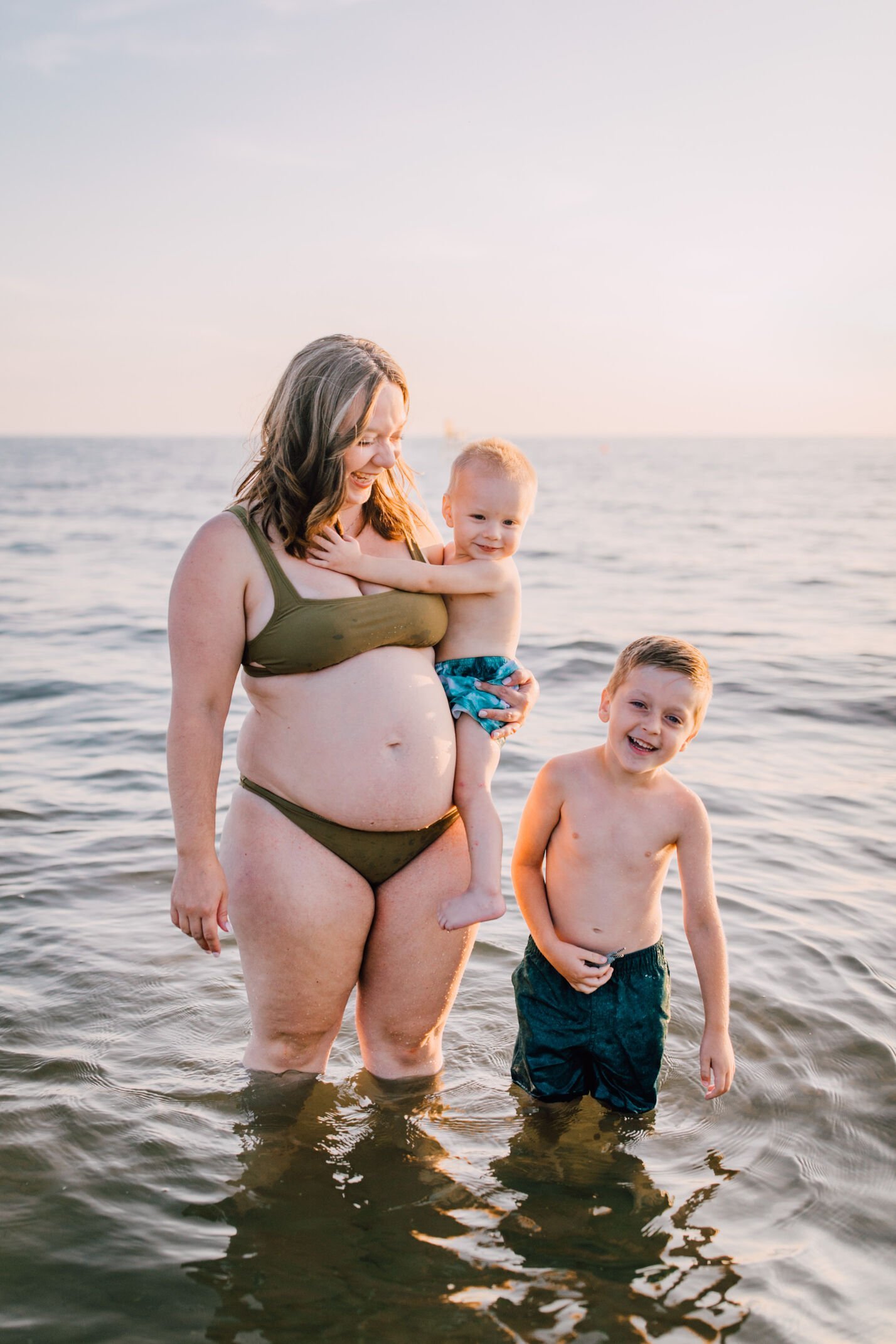  A pregnant mother stands in the water with her sons at southwick beach as they all smile, beach family photos 