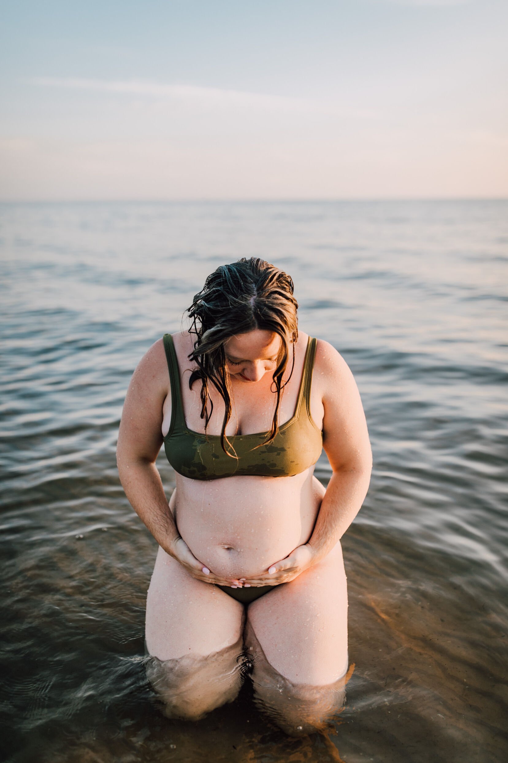  A woman with beach hair sits in the water at southwick beach looking down and holding her pregnant belly, beach maternity photos 
