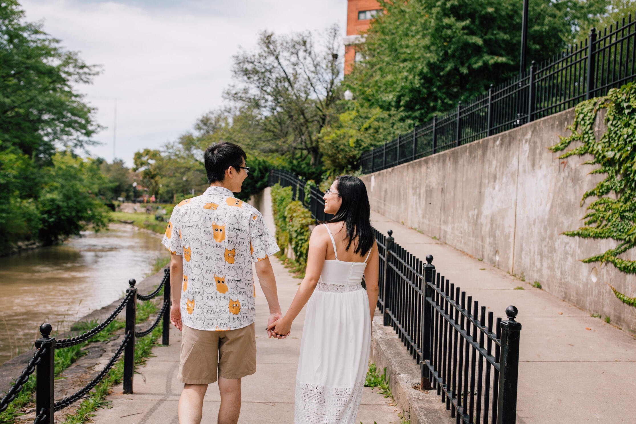  An engaged couple walk hand in hand on a sidewalk next to a creek during their engagement session, Syracuse creekwalk 