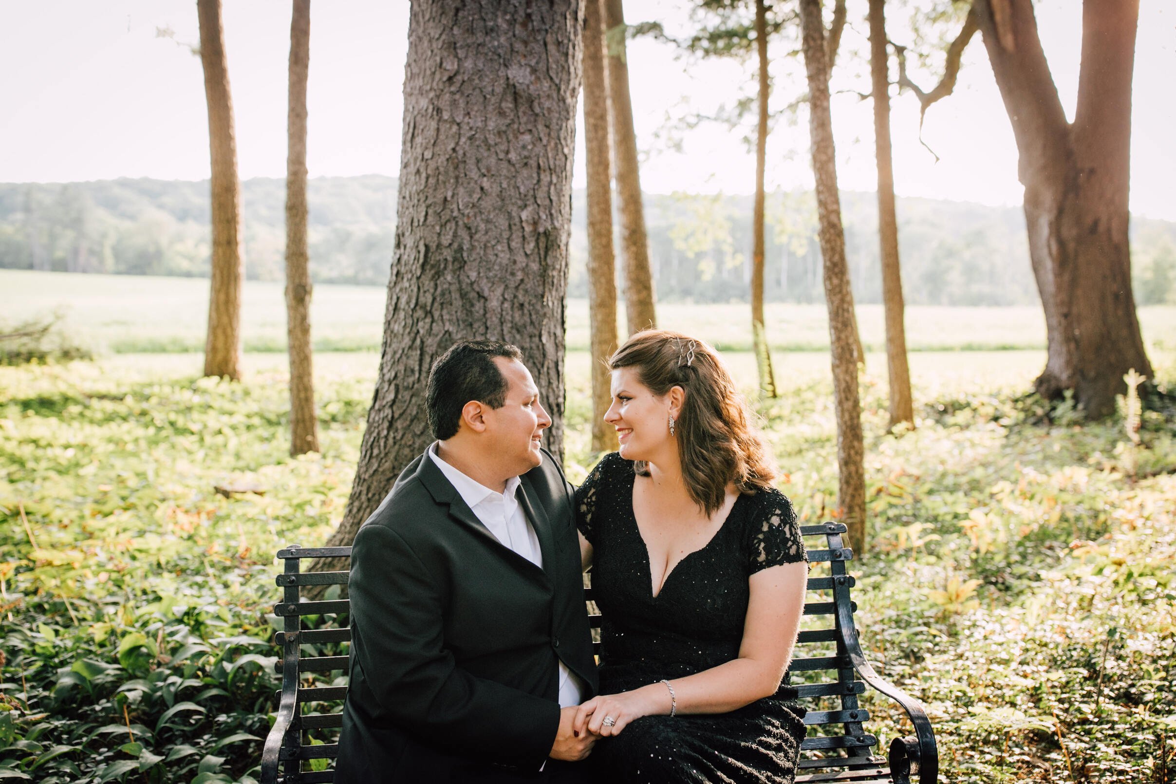  Married couple beam at each other as they sit on a metal bench in a forest at Lorenzo state historic site 