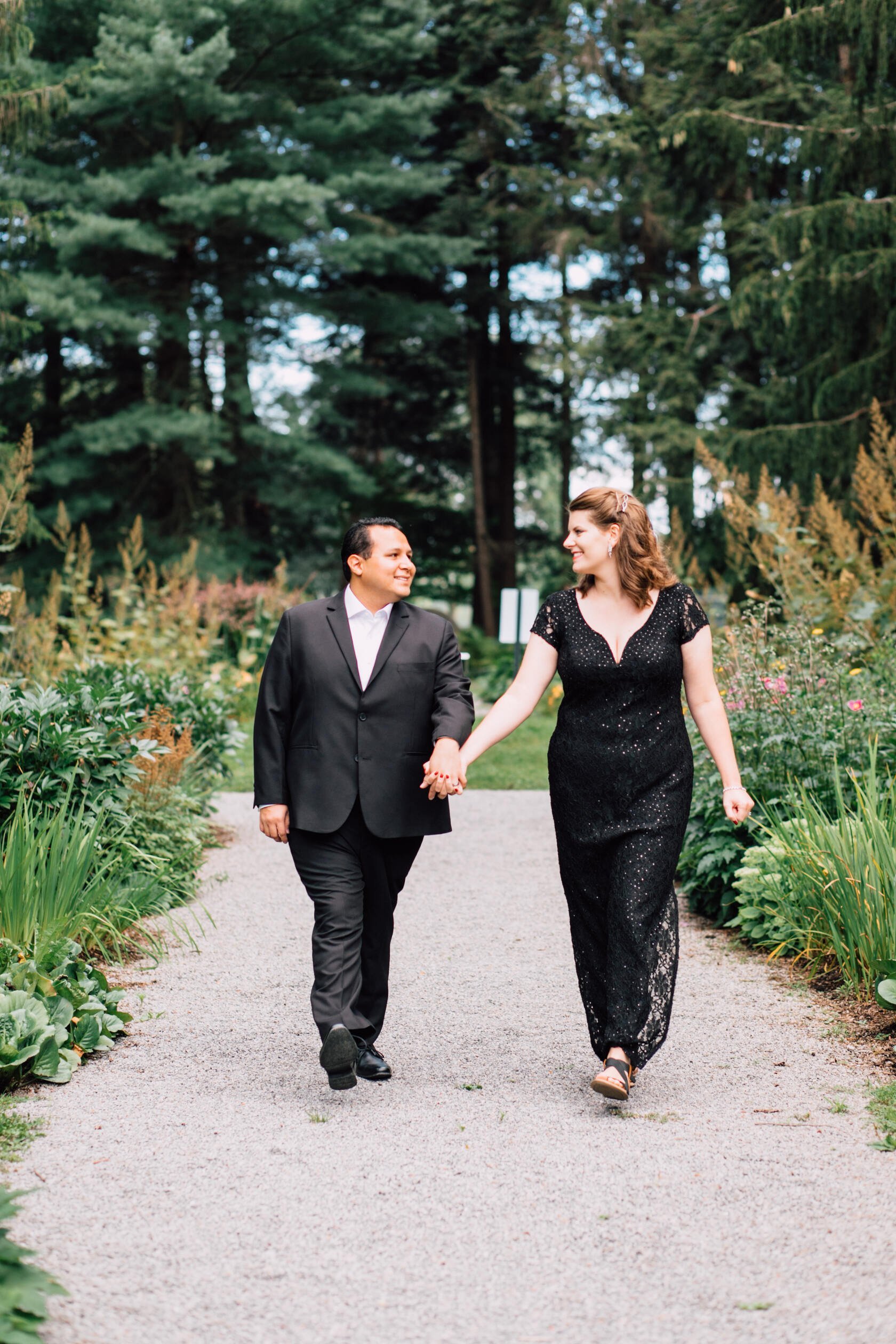  A married couple dressed in all black smile at each other as they walk hand in hand along a gravel path in a garden for anniversary photos 