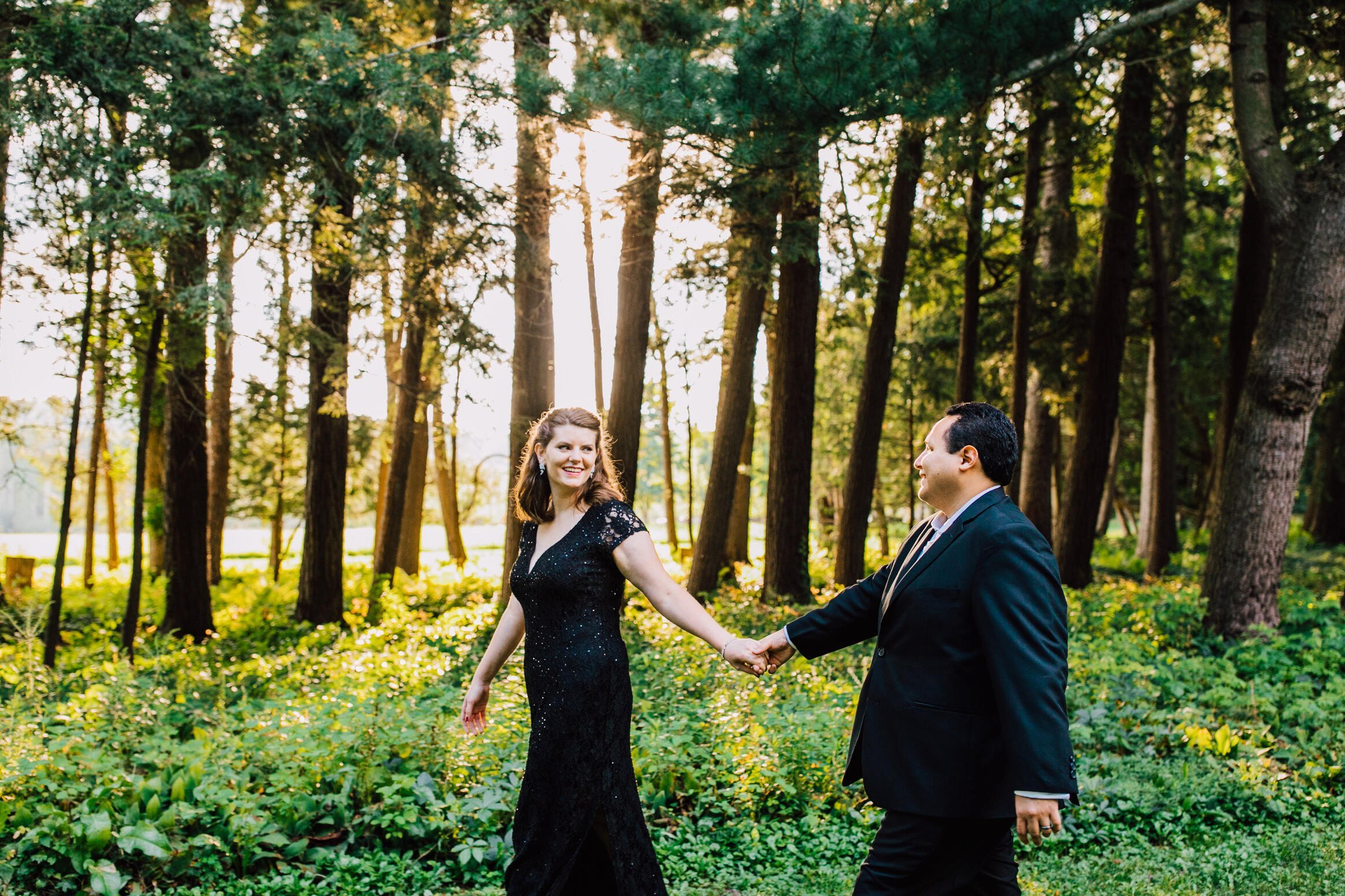  Elizabeth looks back at her husband as they walk hand in hand through woods in Cazenovia, NY for their anniversary 