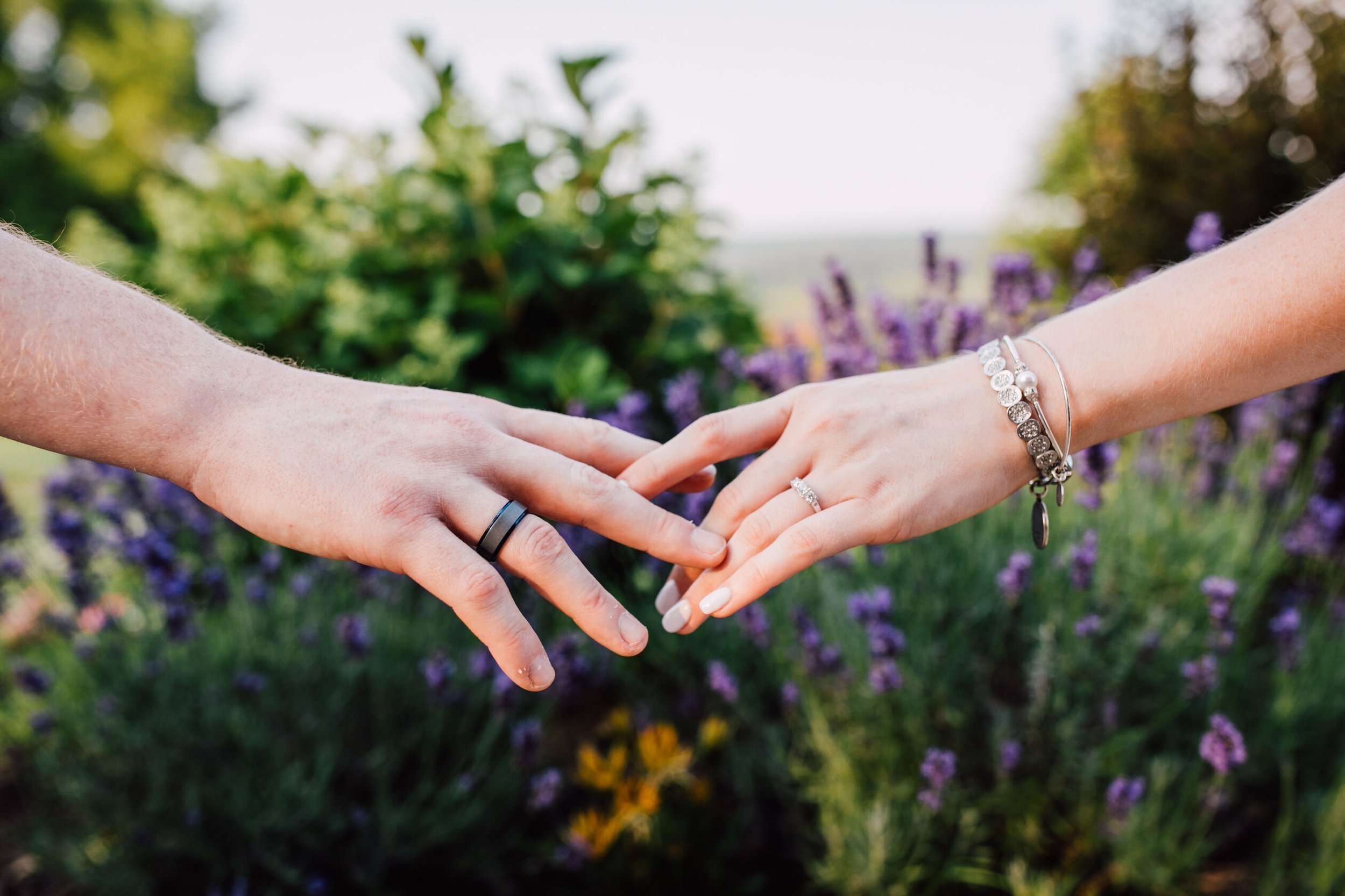  Engaged couple’s hands touch in front of blooming flowers to show their rings for their farm engagement photos 