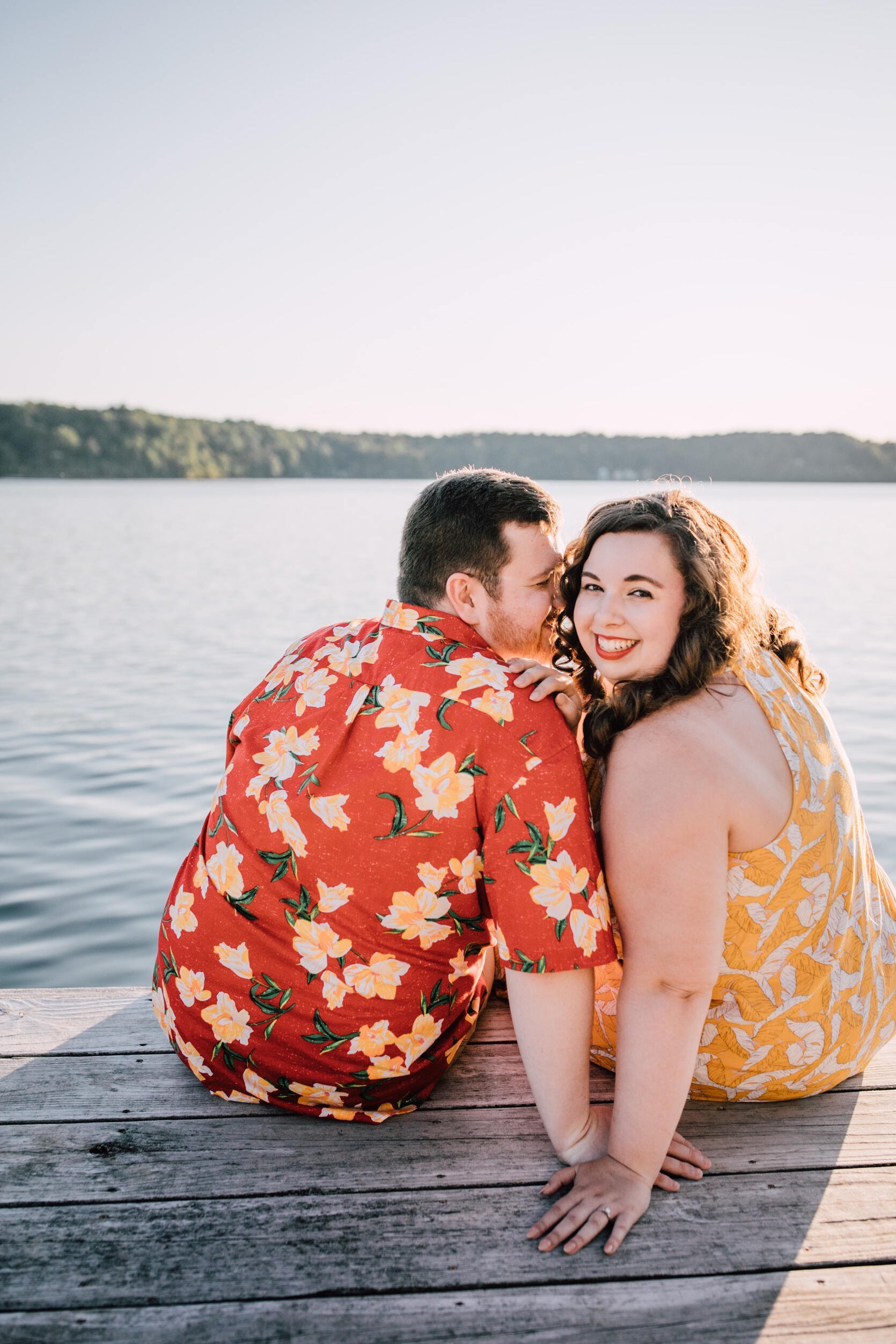  Hanna looks over her shoulder while her fiancé snuggles in on a dock in from of Lake Ontario for their sunset engagement photos 