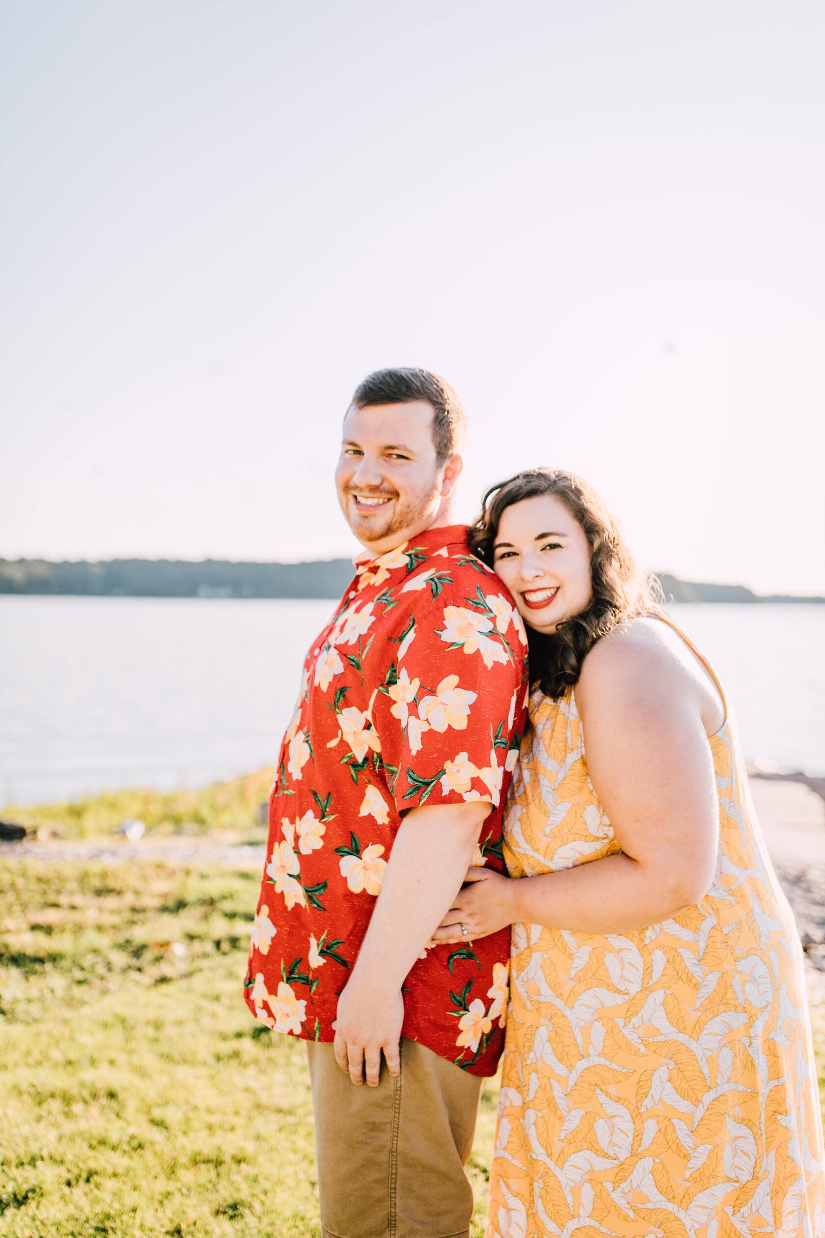  Hanna rests her head on her fiancé’s shoulder while they smile together for their lake engagement photos 