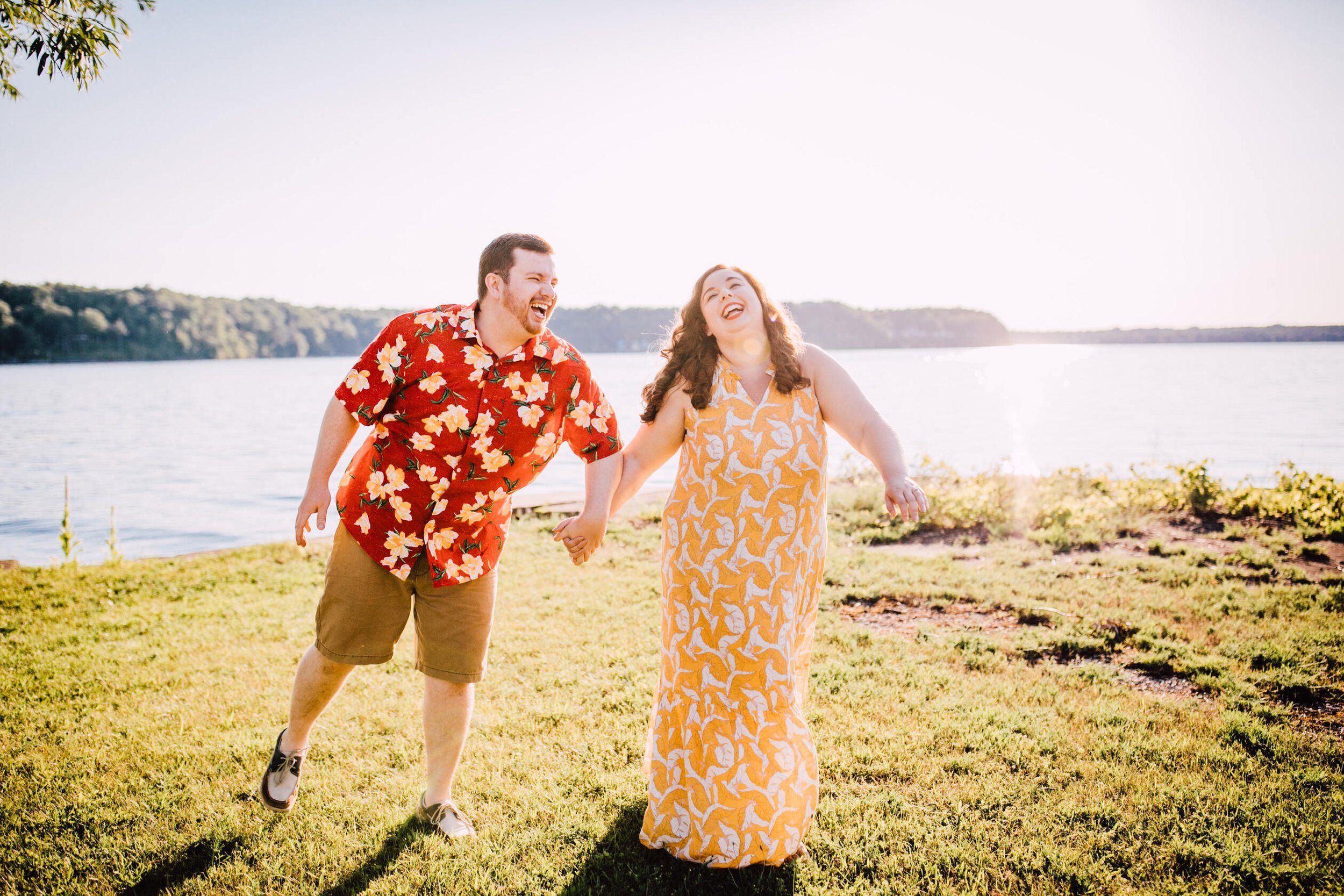  Engaged couple laugh together while walking and holding hands in a grassy area in front of Lake Ontario for their sunset engagement photos 