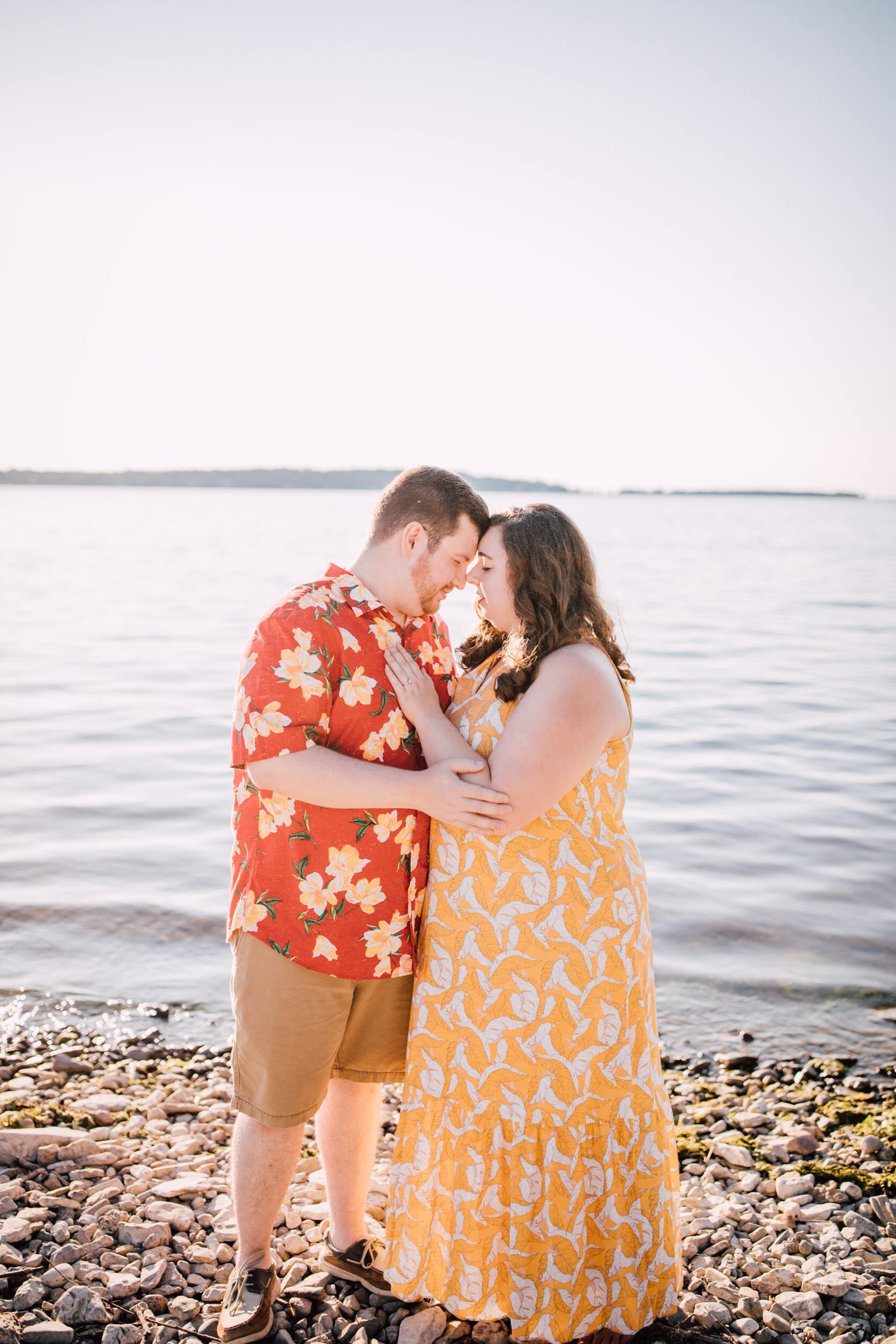  Hanna rests her hand on her fiancé’s chest as they lean in to touch foreheads while standing on a rocky beach for lakefront photography 