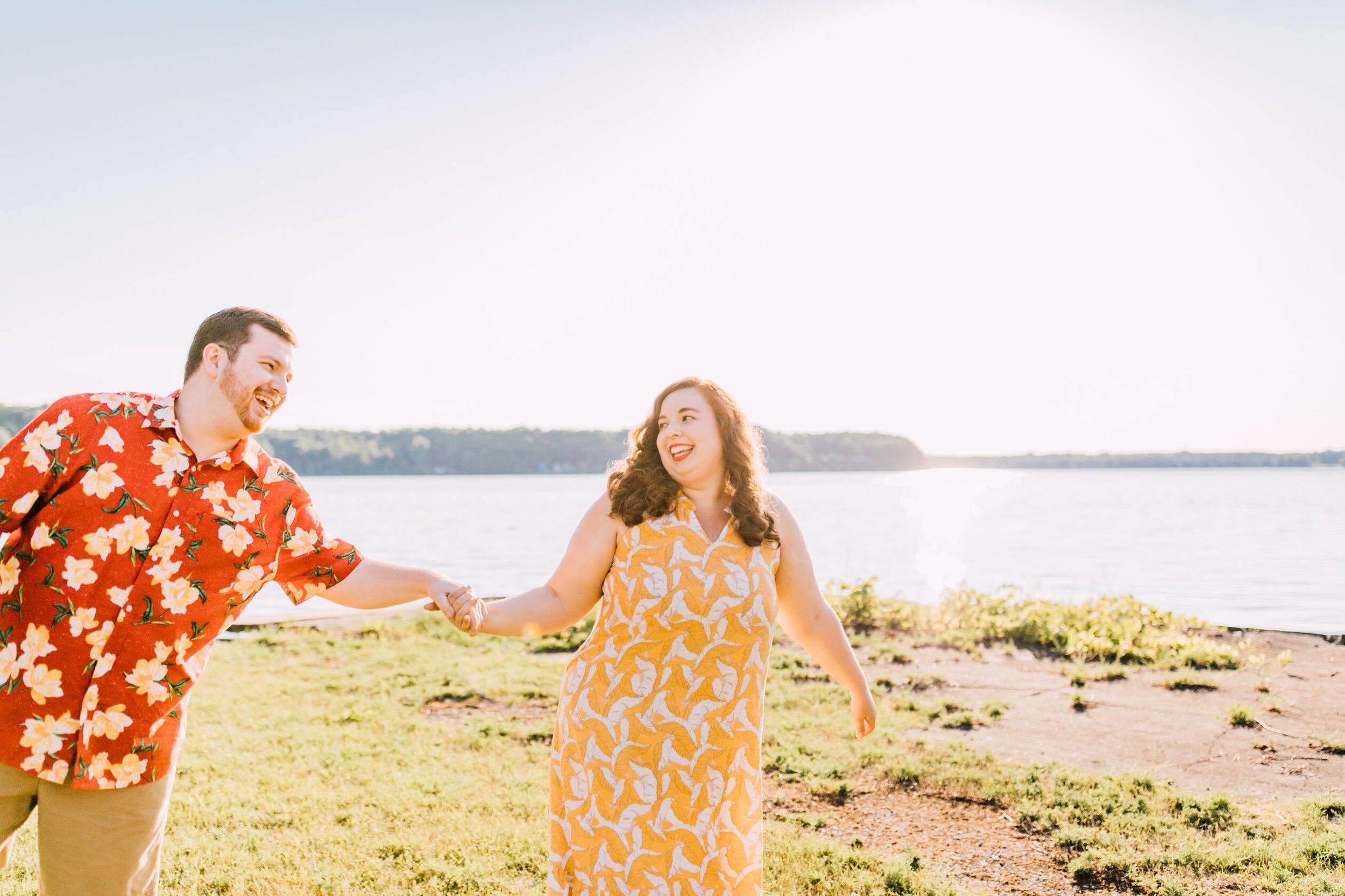  Hanna and Brandon’s sunset engagement photos show off their personality perfectly. Here, they hold hands while laughing together in a grassy area in front of Lake Ontario 