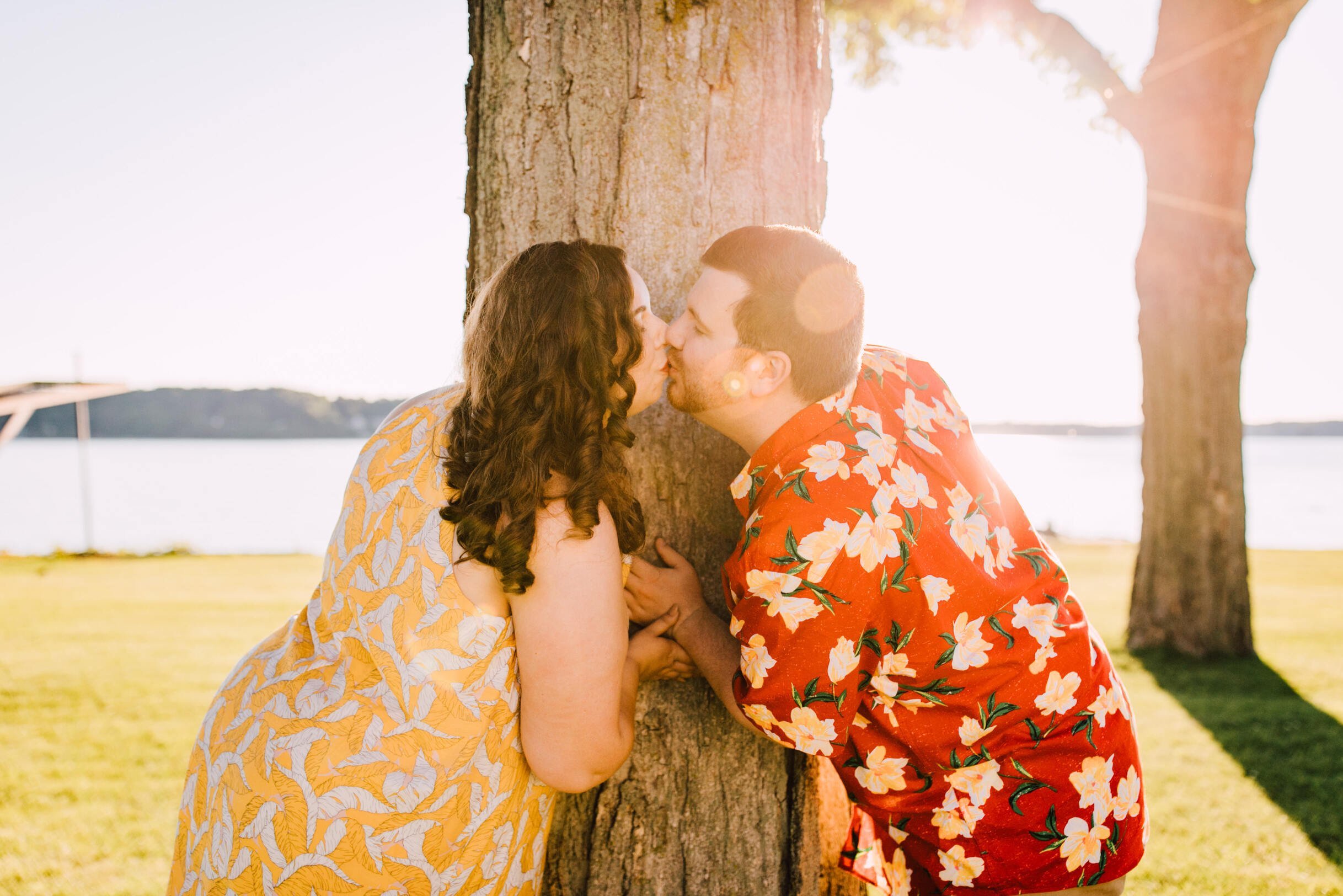  Engaged couple kiss while wrapped around a tree in a grassy area in front of the lake making for stunning lakefront photography 