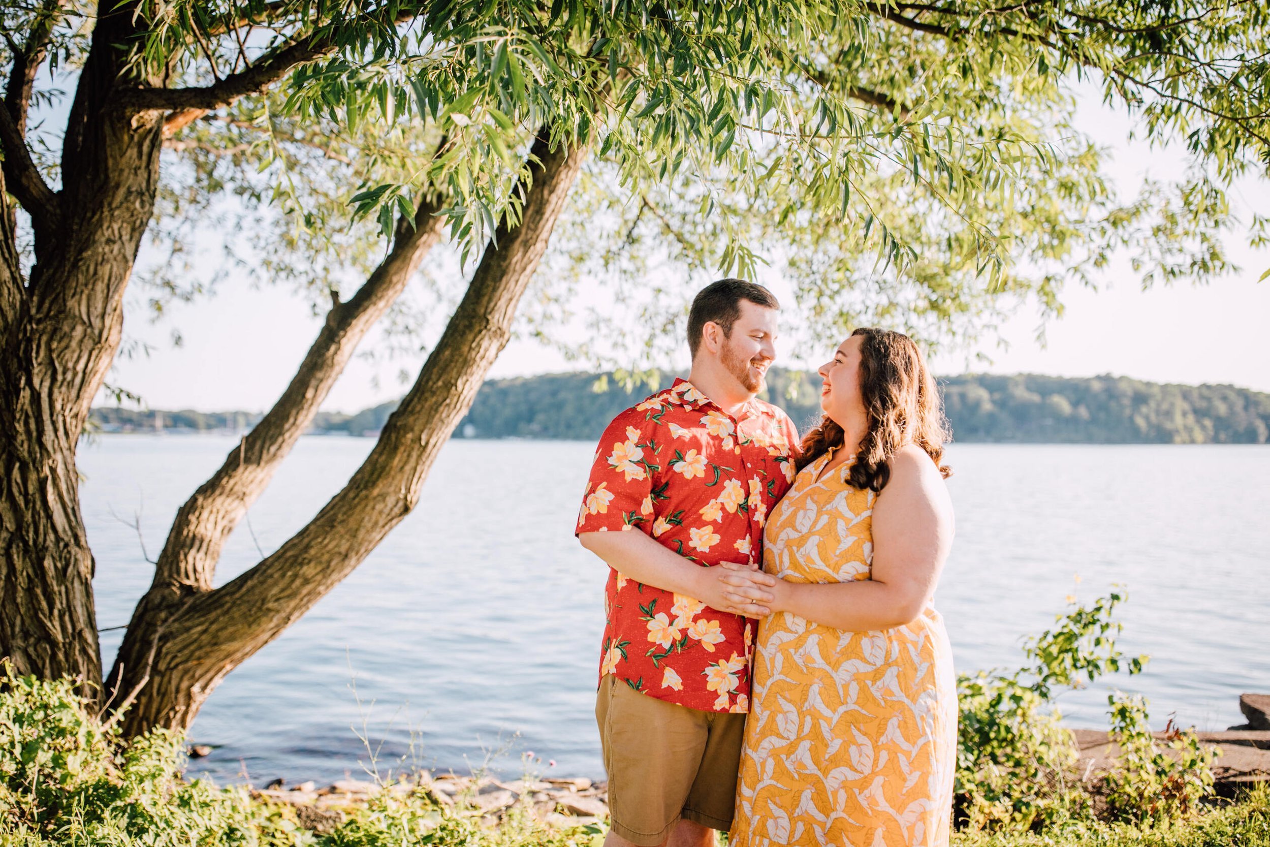  Engaged couple stand close to each other while smiling at each other wearing colorful engagement outfits in a grassy area in front of a Great Lake 