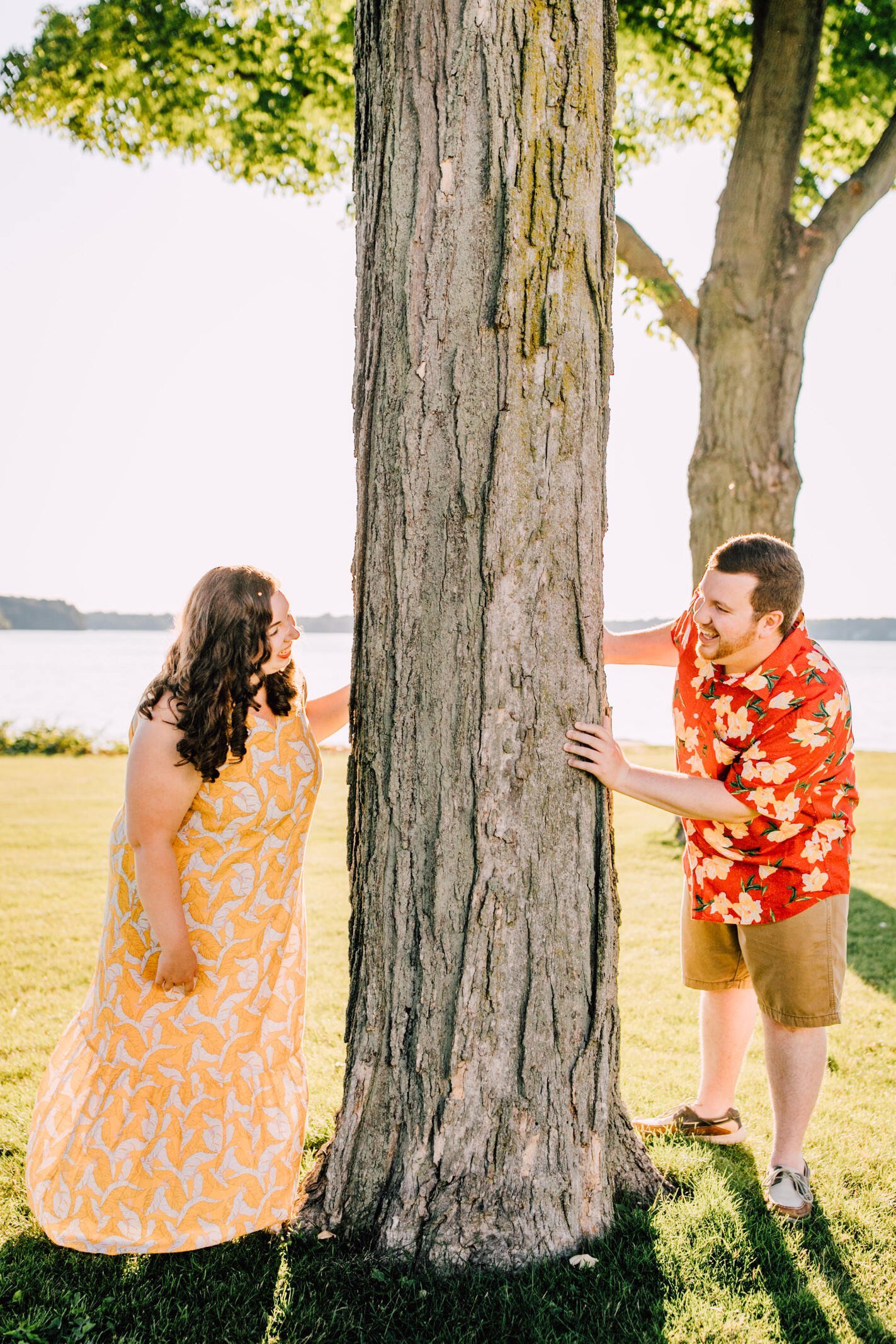  Hanna and Brandon smile at each other from opposite sides of a tree during sunset engagement photos in a grassy area with the lake at the horizon 
