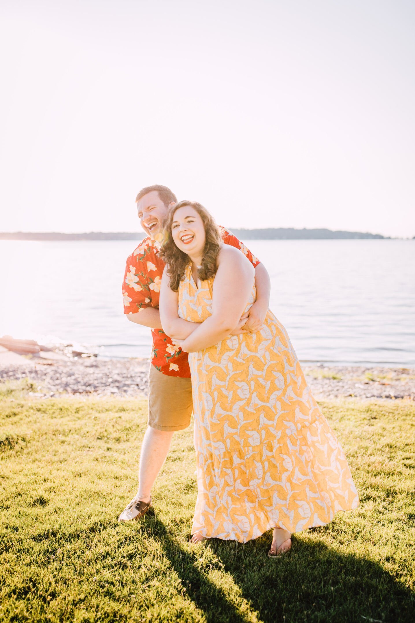  Brandon laughs with his arms wrapped around his fiancé as they dance in a grassy area in front of the lake for sunset engagement photos 