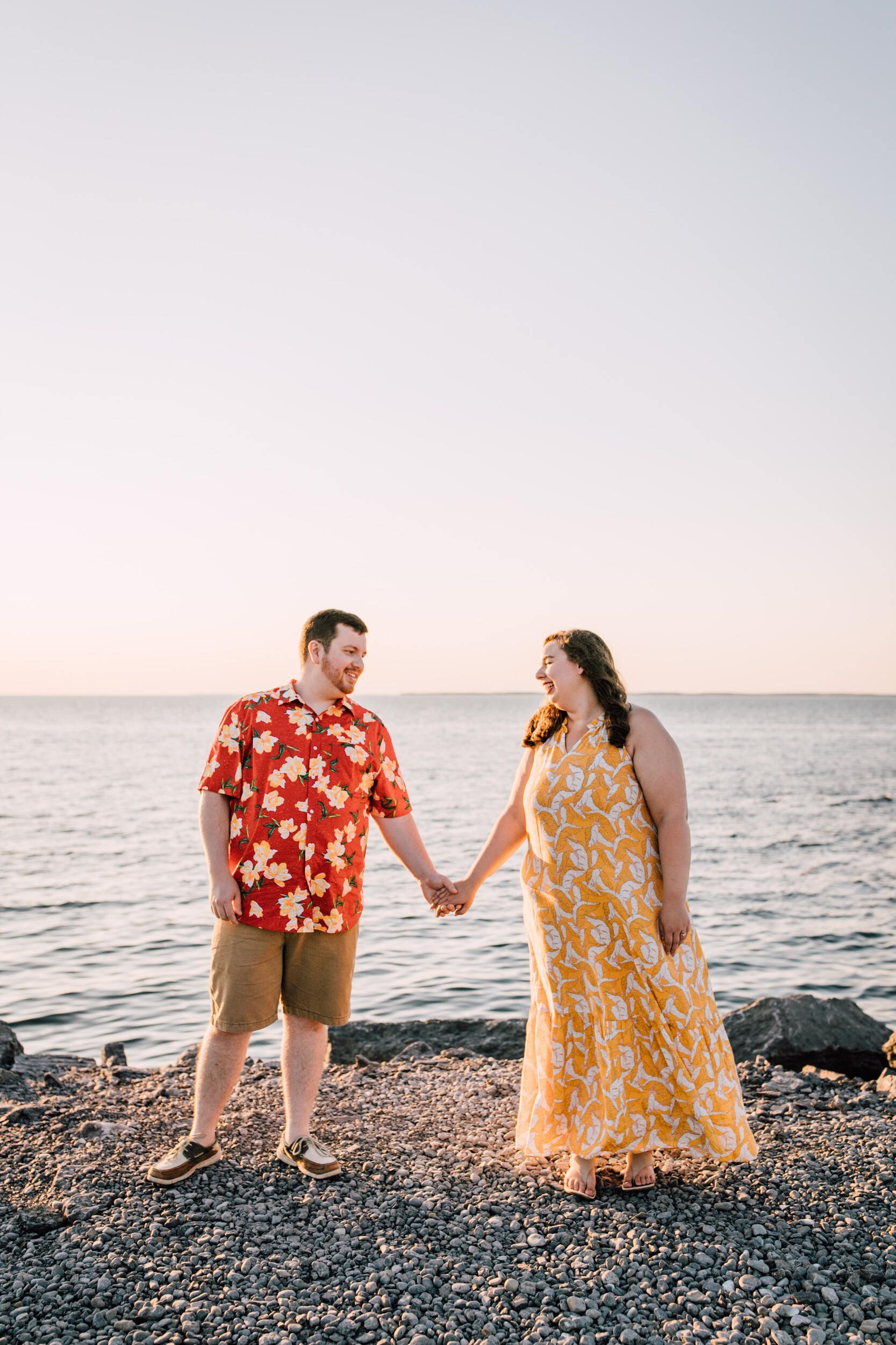  Lakefront photography is at its peak for Brandon and Hanna’s engagement session. Here, they hold hands while smiling at each other on a rocky beach in front of Lake Ontario 