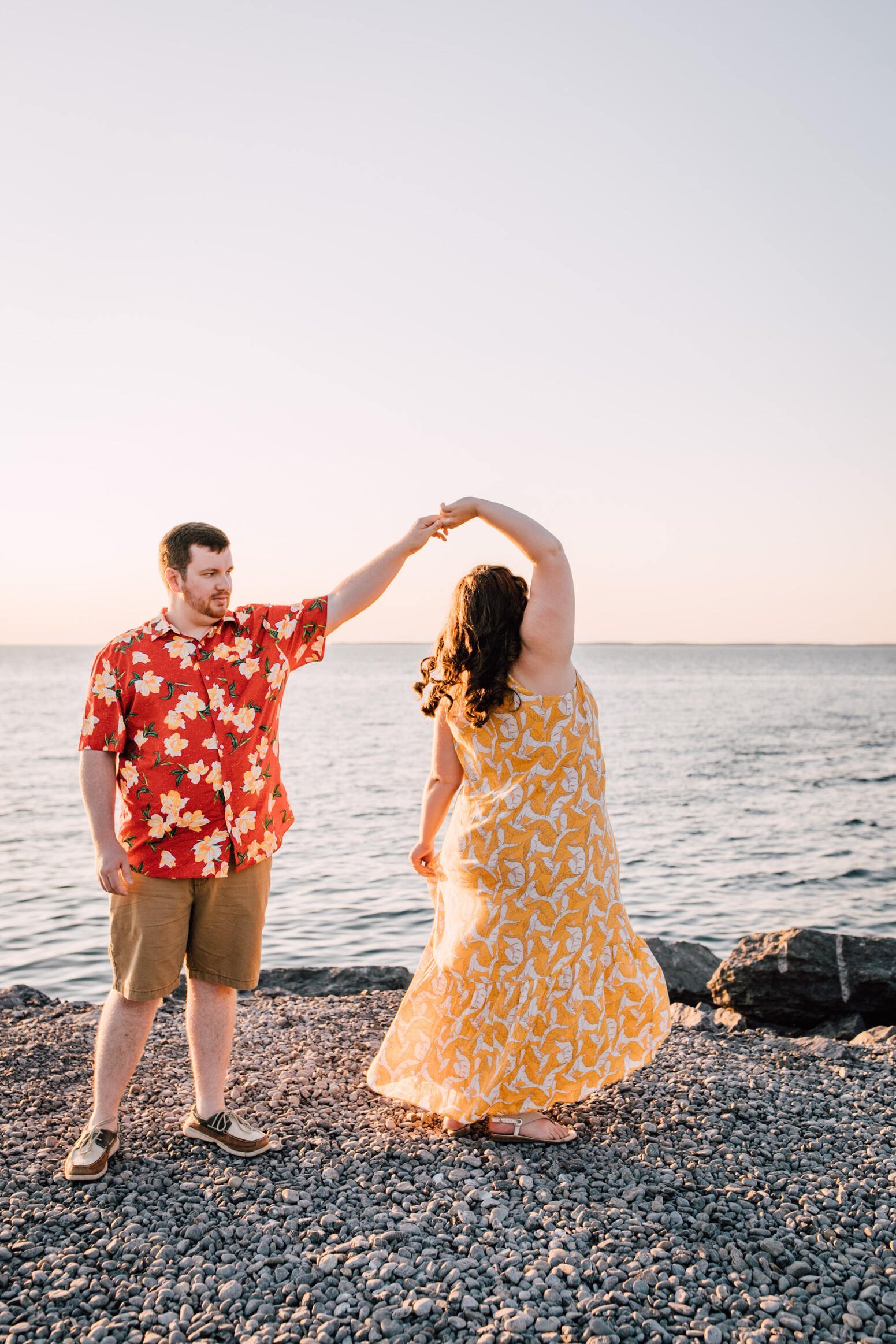  Brandon spins his fiancé as they dance on a rocky beach on the shore of Lake Ontario for their lake engagement photos 