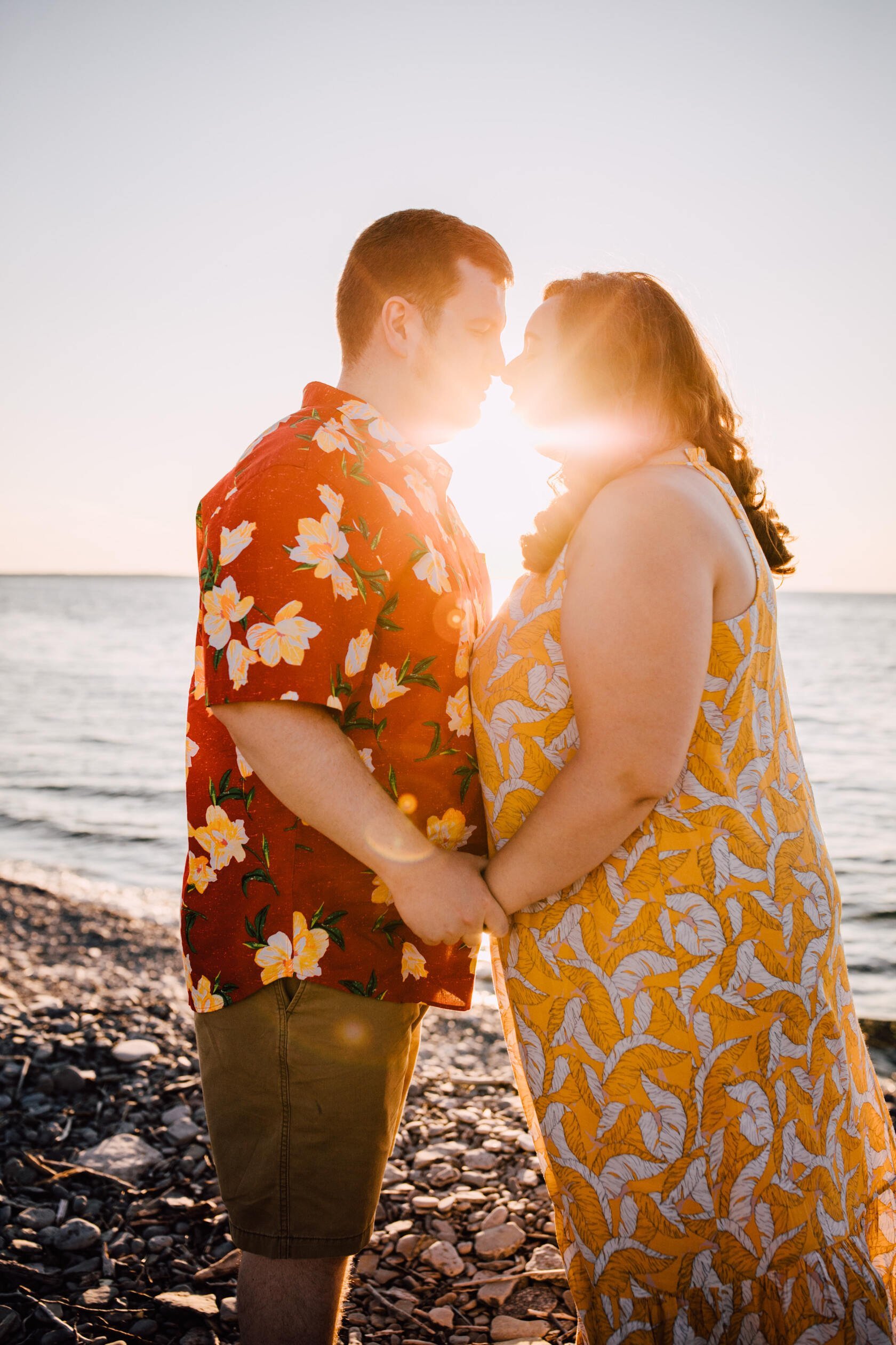  Engaged couple faces each other as they lean in close with the sun shining through behind them as it sets on the rocky beach shore of lake Ontario making for gorgeous sunset engagement photos 