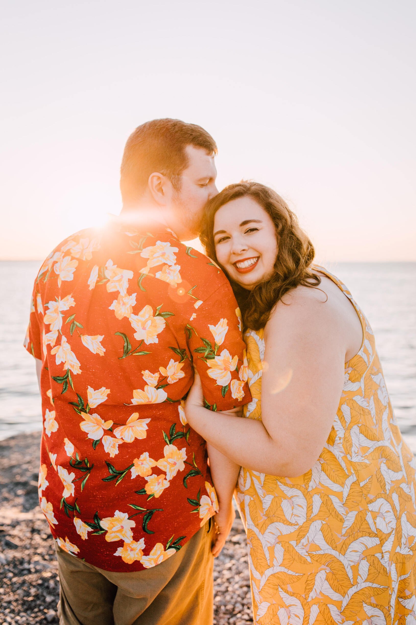  Brandon kisses the top of his fiancé’s head as she gazes over her shoulder with a wide smile. This example of lakefront photography is taken on the rocky shore of Lake Ontario 